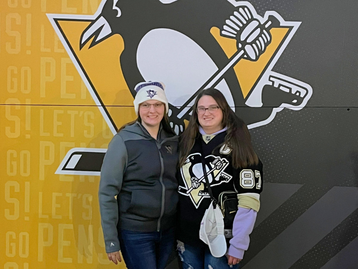 Two fans pose in front of a Pittsburgh Penguins mural; one wears a Penguins jersey, the other a gray jacket and white knit hat, both smiling.