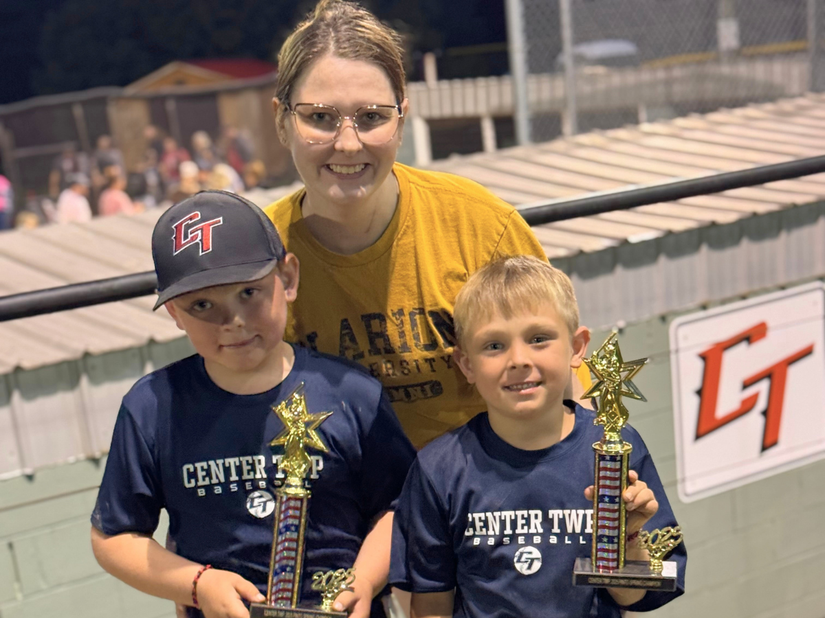 Smiling woman stands behind two boys in navy shirts, each holding a trophy at a baseball field.