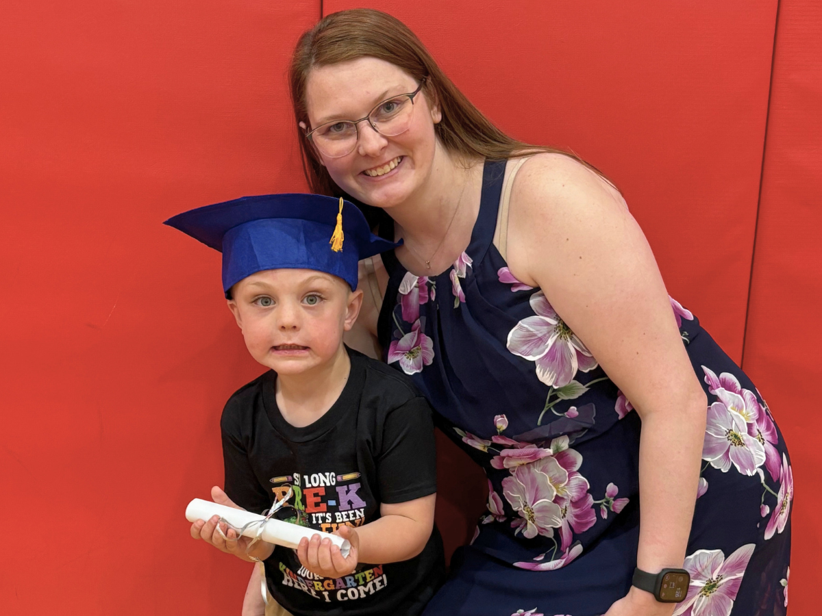 A smiling woman in a floral dress poses with a young child wearing a blue graduation cap and holding a rolled diploma against a red wall.
