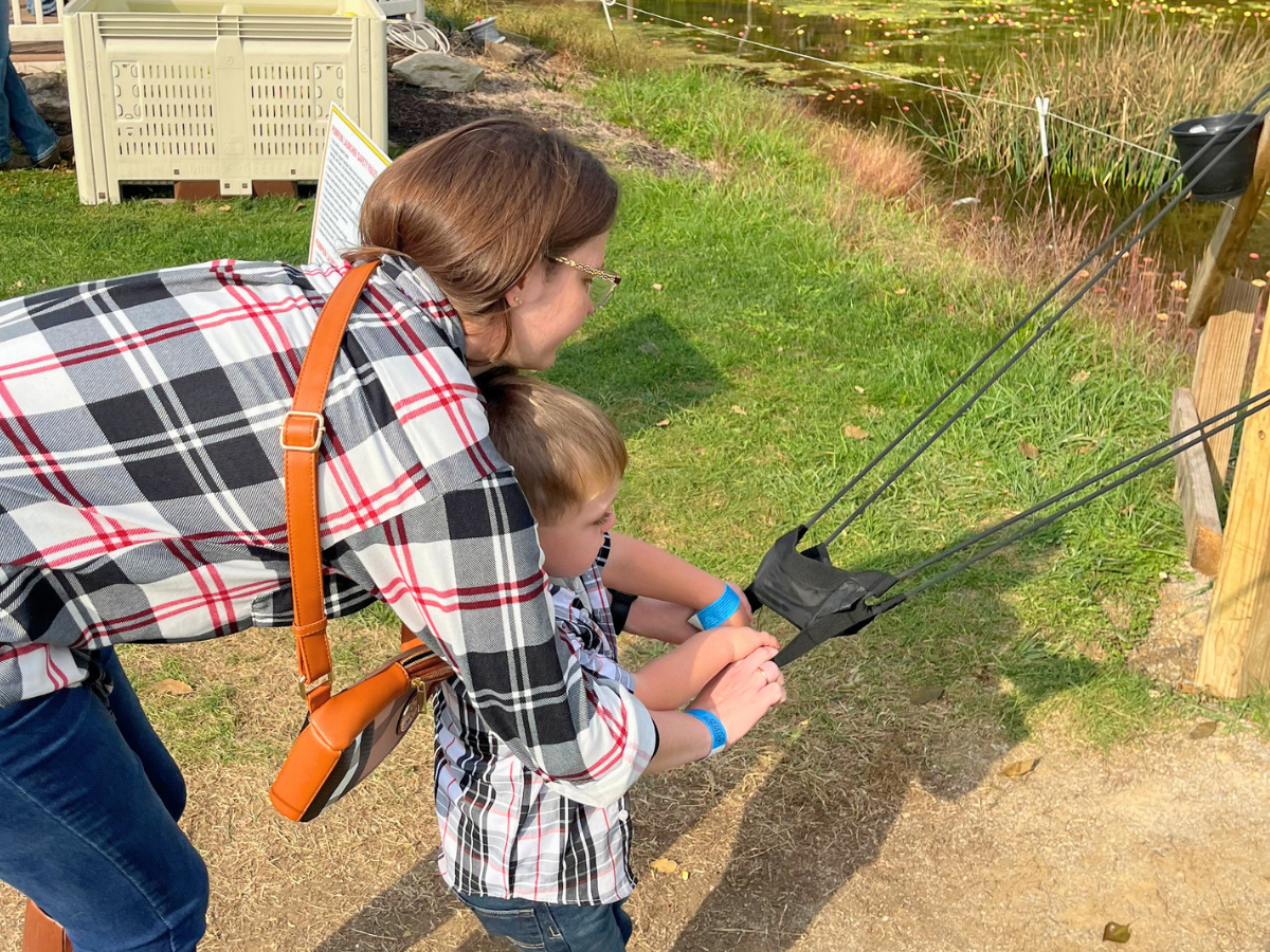 Woman helping a young boy hold a harness connected to several cords outdoors near a grassy area and a pond, during a park demo or activity.