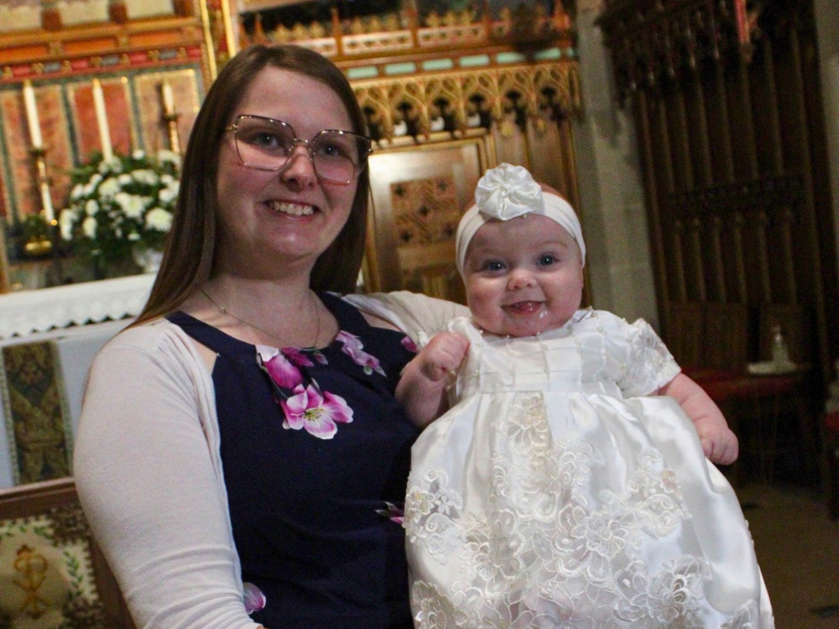 Smiling woman in glasses holding a baby girl in a white lace dress inside a church.
