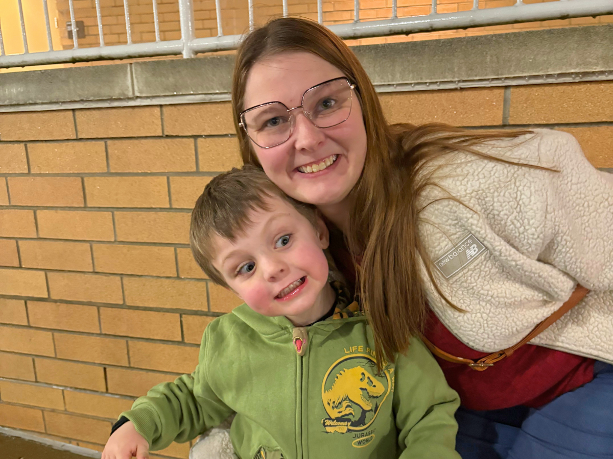 Smiling woman with glasses leaning over a smiling boy in a green hoodie against a brick wall in an indoor setting.