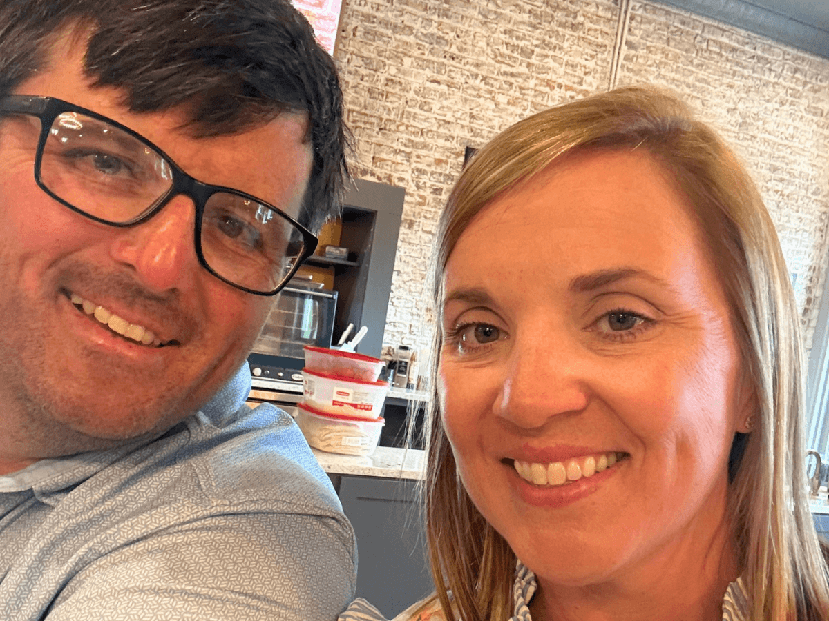 Close-up selfie of a man wearing glasses and a woman, both smiling, in a kitchen with a brick wall background.