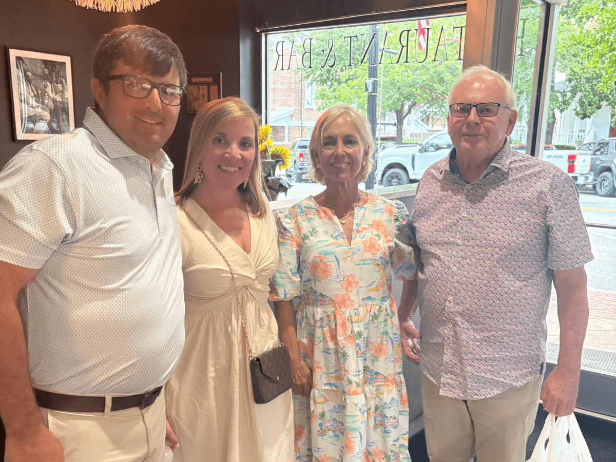 Four adults posing together in a cafe, smiling at the camera by a large window.