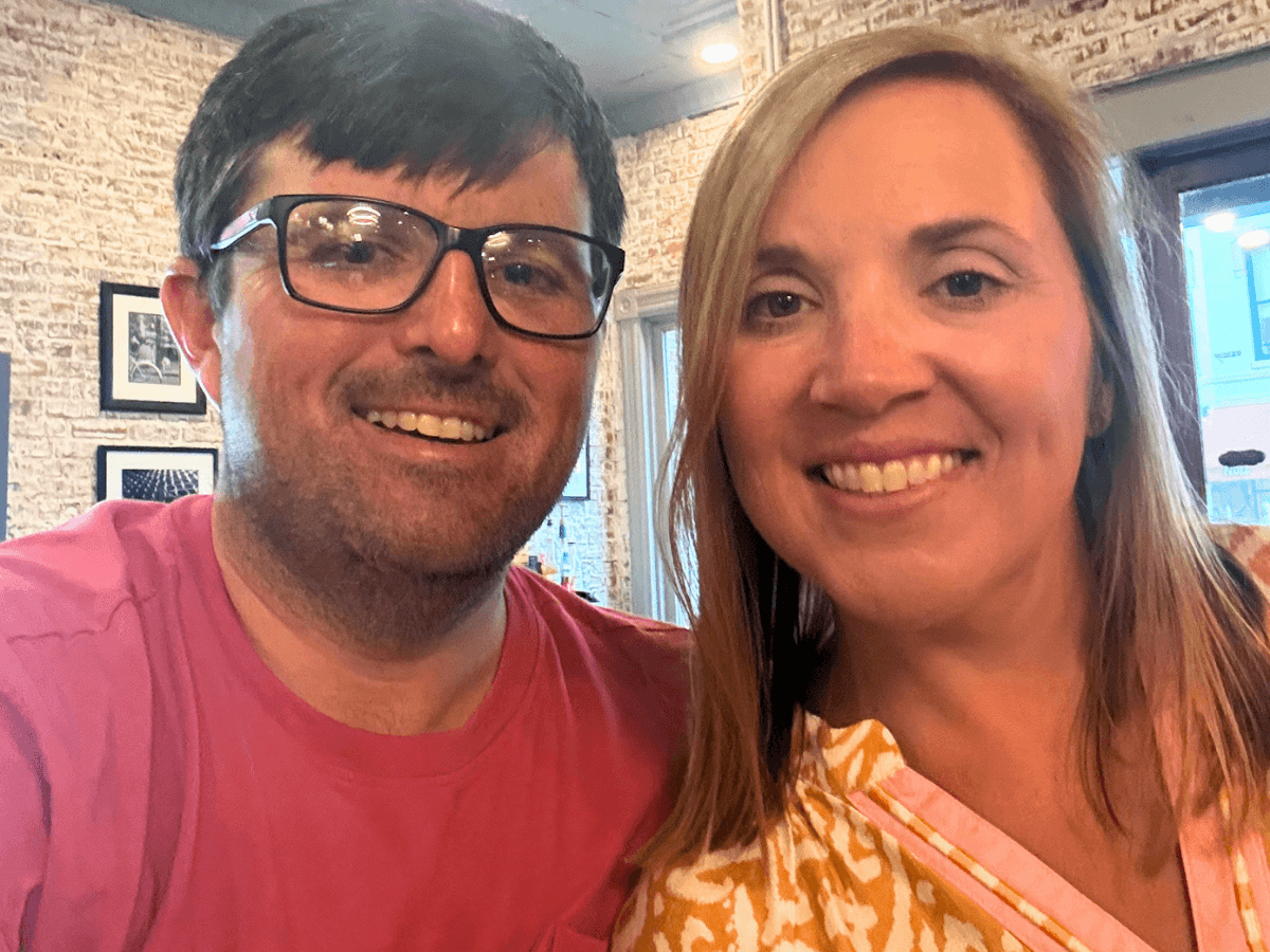 Smiling man with glasses and a pink shirt next to a smiling woman with blonde hair in a brick-walled cafe.