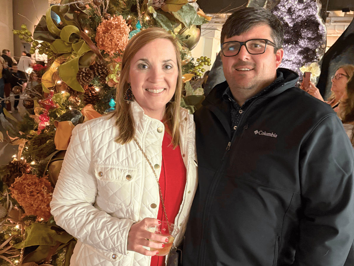 A smiling couple posing in front of a decorated Christmas tree at a holiday event; woman in white jacket holding a drink, man in a black Columbia jacket beside her.