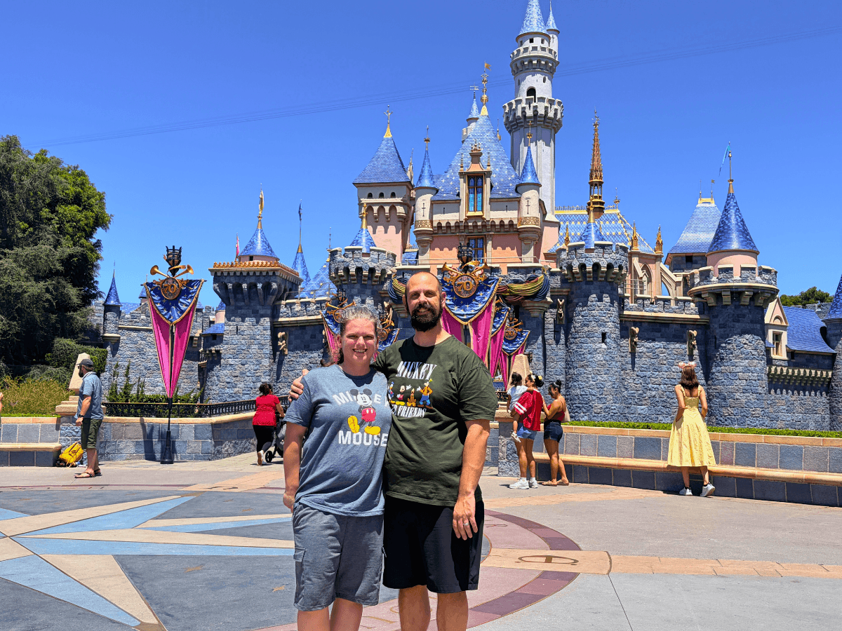 Couple posing and smiling in front of a blue-roofed fairy-tale castle at a theme park, with banners on the walls.
