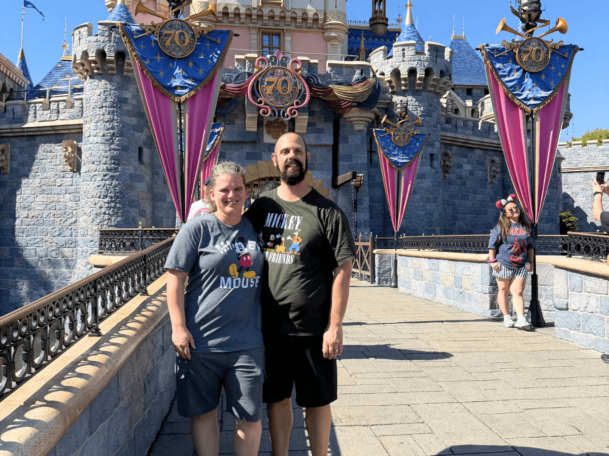 Smiling couple standing on a sunlit walkway in front of Disneyland's castle adorned with blue and pink 70th anniversary banners.