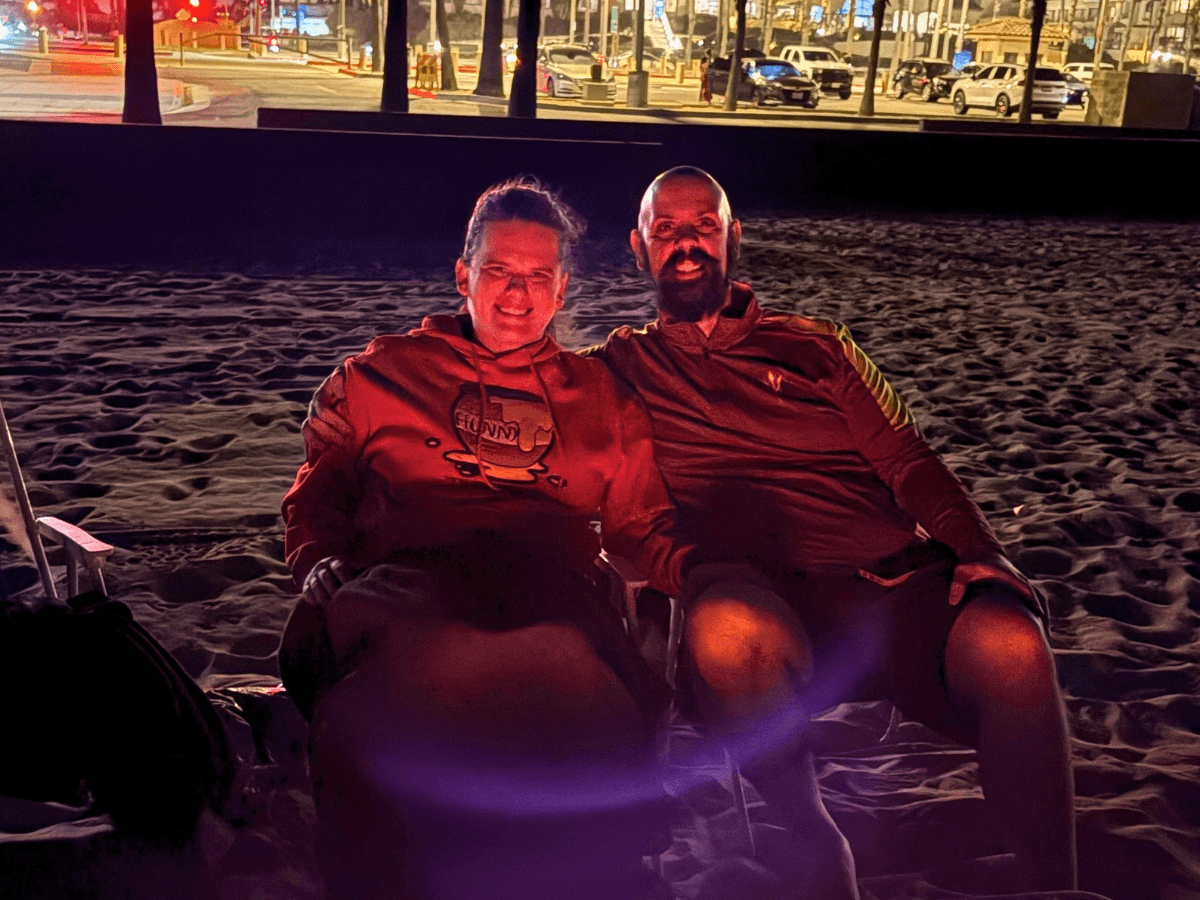 Two people sit close together on a sandy beach at night, smiling at the camera with a city street and lights in the background, wearing casual long-sleeve tops.