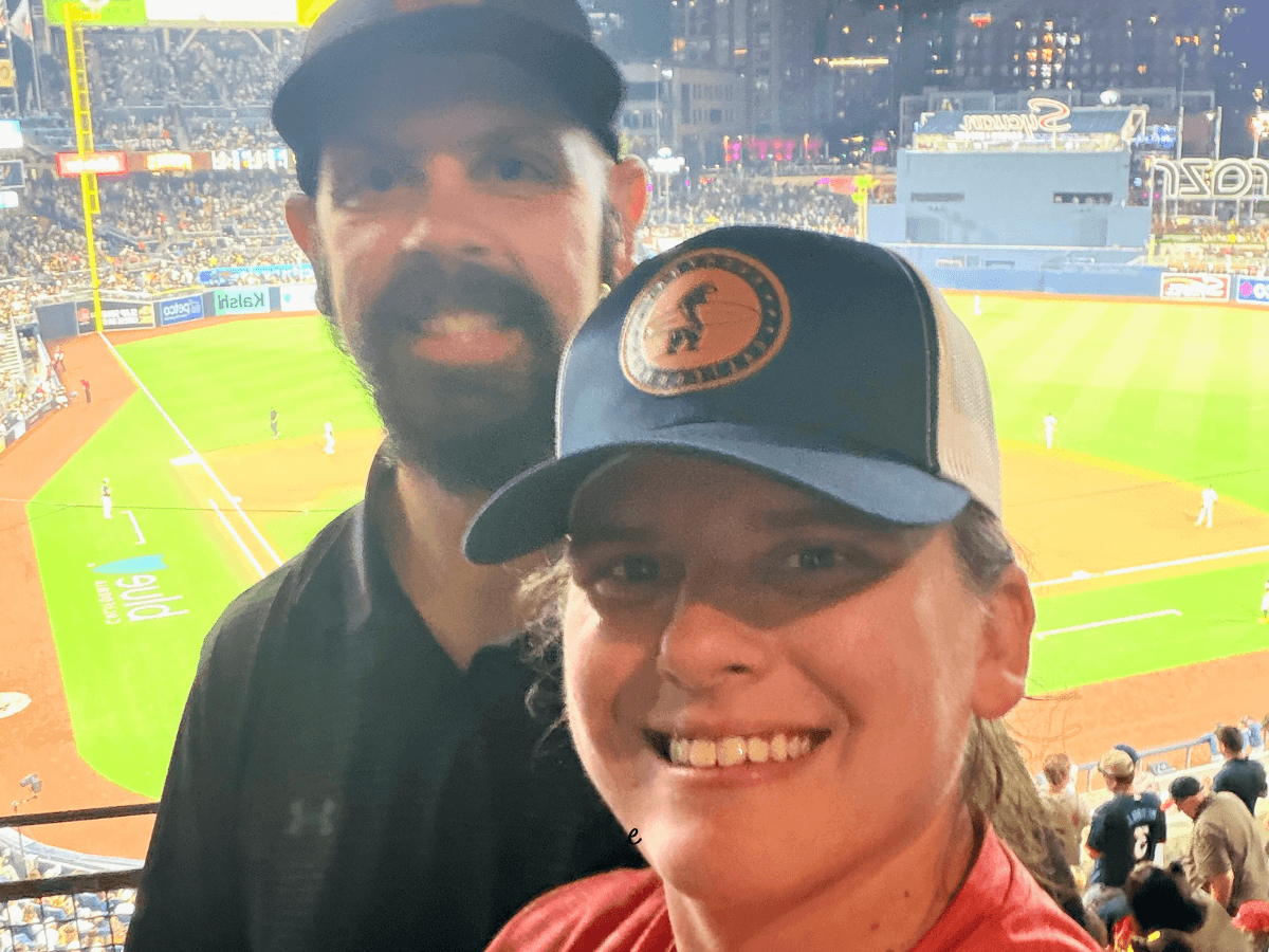 Two fans pose for a selfie at a baseball stadium, with the field and crowd behind them at night.