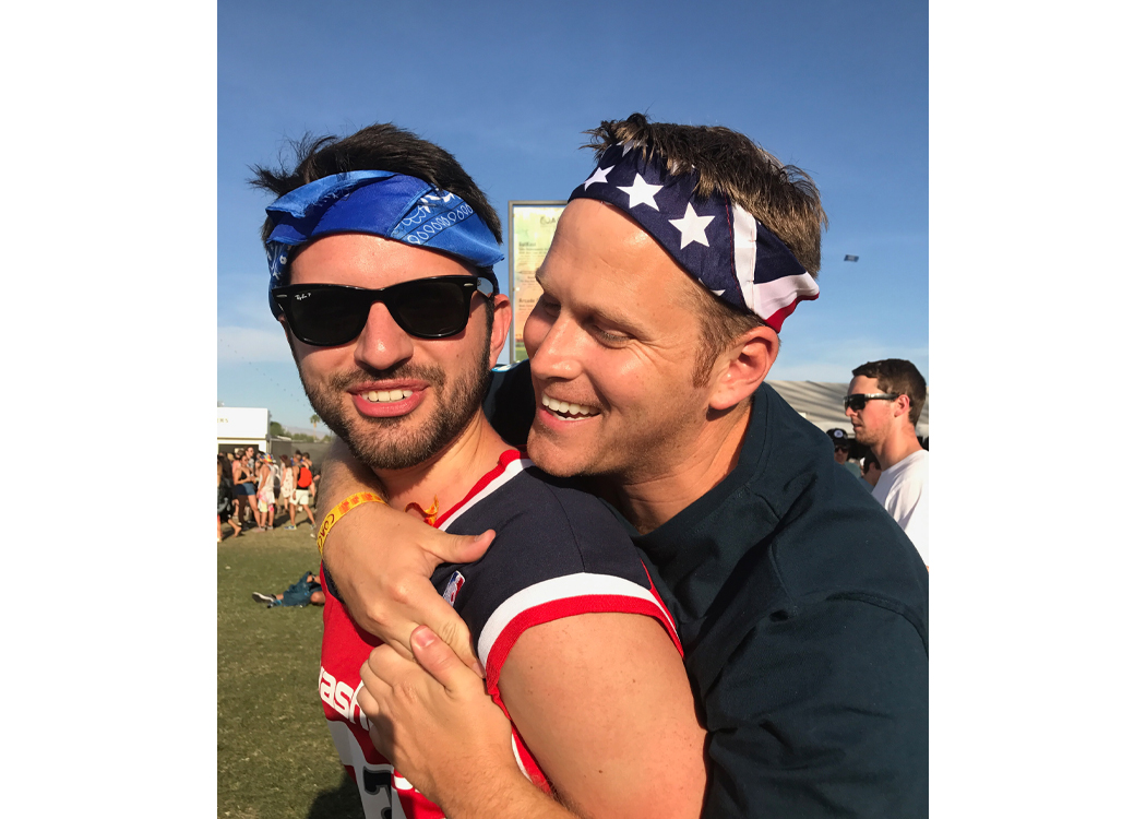 Two friends at an outdoor event hug; one in a red athletic shirt with a blue headband and sunglasses, the other in a stars-and-stripes bandana smiles while hugging from behind.
