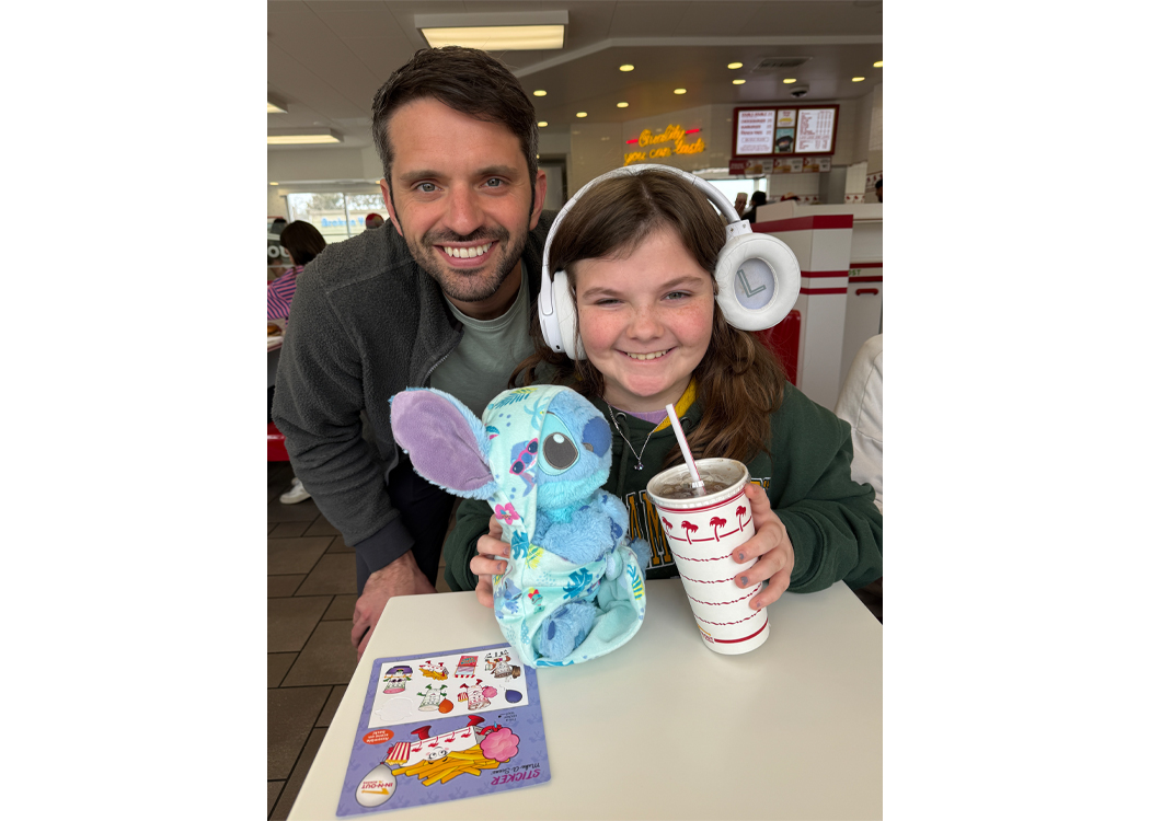 Smiling man leans over a table next to a girl wearing large white headphones, holding a blue Stitch plush toy and a tall cup with a straw at a casual restaurant.