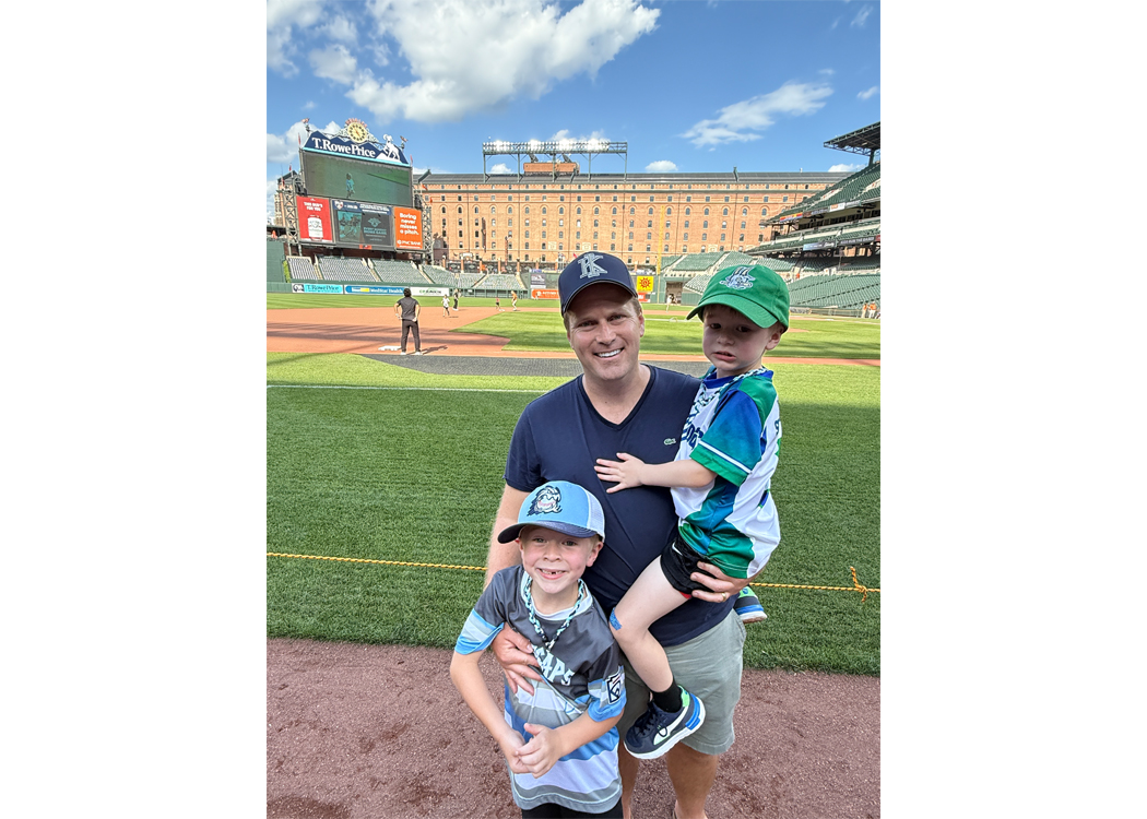 Father with two young sons posing on the baseball field, smiling at the camera with stands and scoreboard in the background.