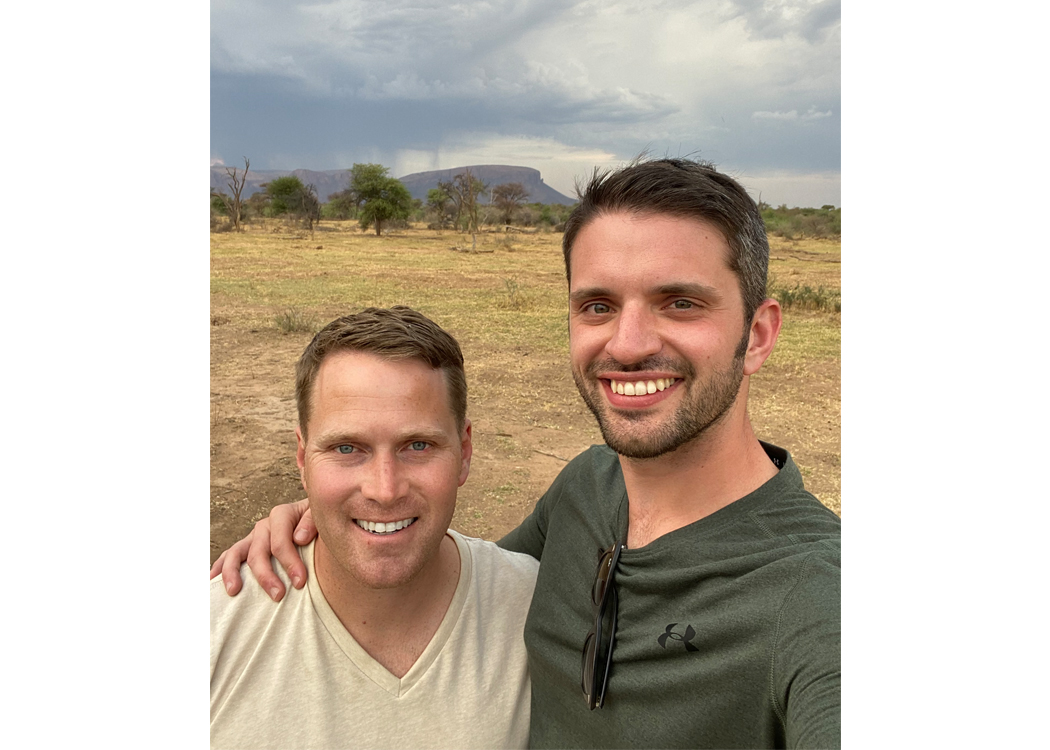Two men smile for a selfie in a dry, open landscape with sparse trees and distant hills under a cloudy sky.Two friends pose close together for a selfie outdoors in a savanna-like field; one in a beige shirt, the other in a green shirt with sunglasses hanging from it. Selfies of two men standing arm-in-arm, smiling at the camera in a sunny, arid landscape with scattered trees. A cheerful outdoor photo of two men in casual shirts, posing for a selfie in a rural, brushy plain with distant hills.