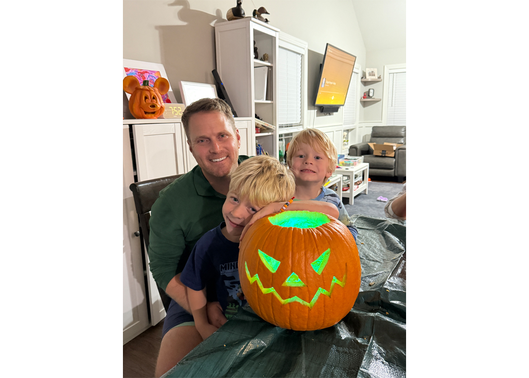 Father with two young boys posing behind a glowing jack-o'-lantern on a table indoors during pumpkin-carving night.