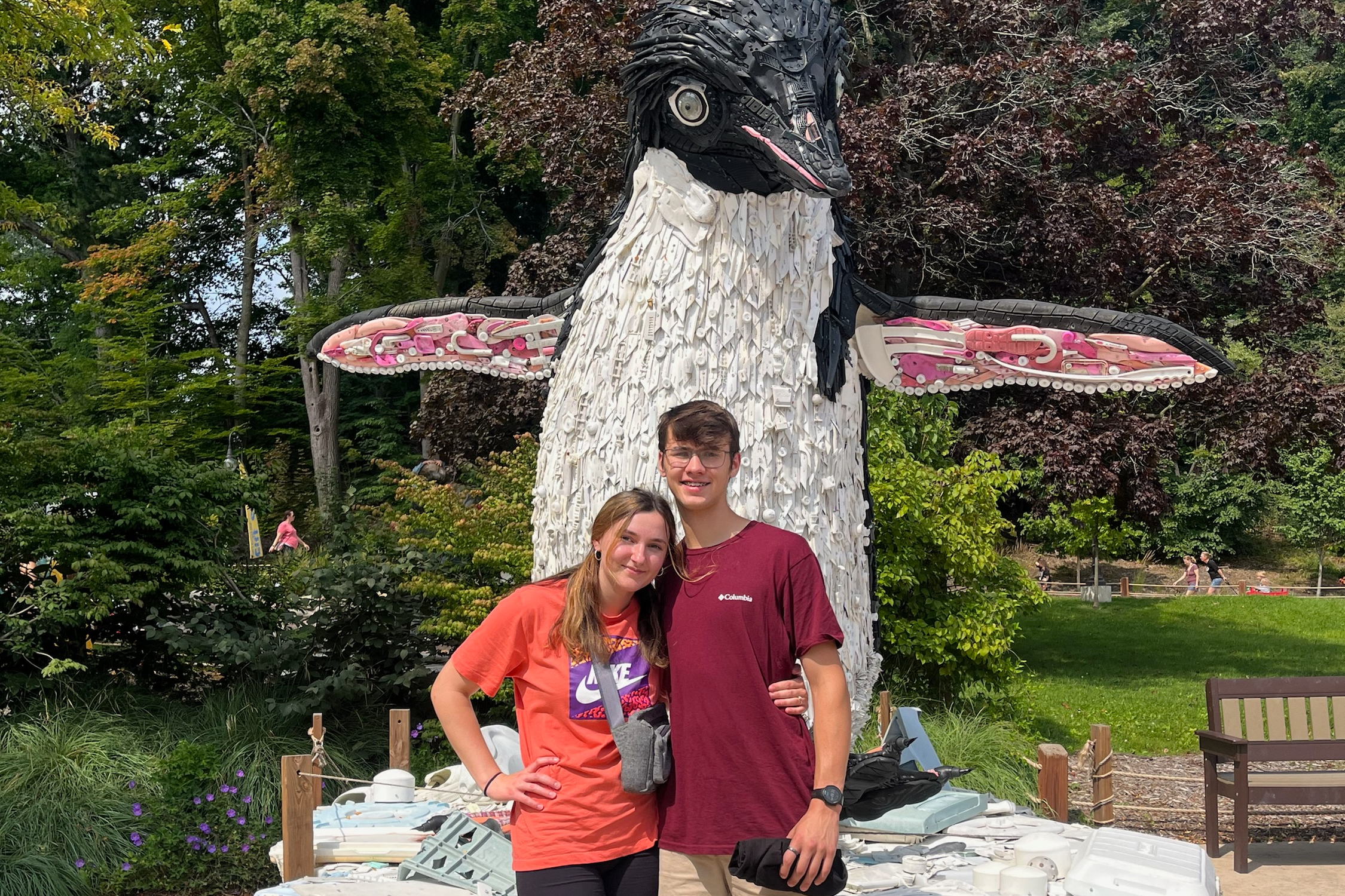 Two teens pose in front of a tall dragon sculpture made from white and black recycled materials with pink wing tips, in a park setting.