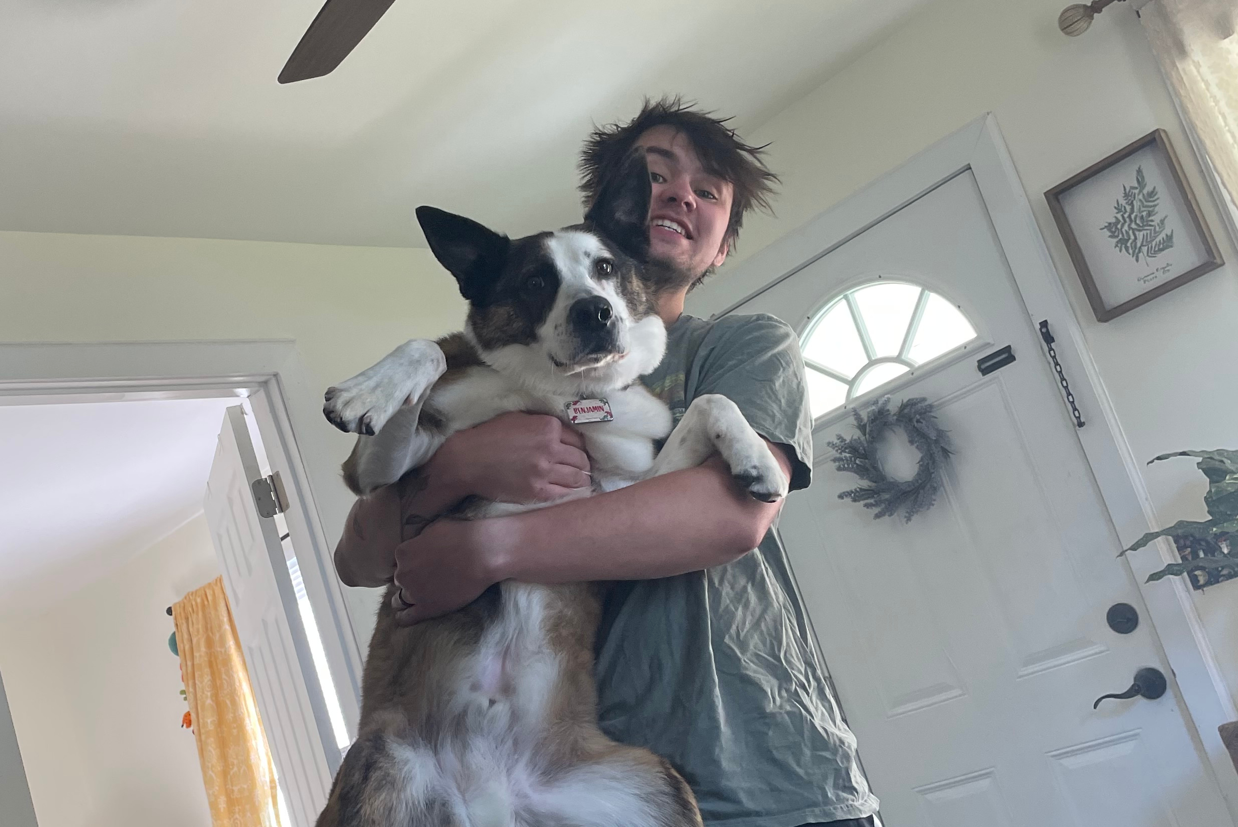 Person smiling while holding a large mixed‑breed dog indoors near a front door.