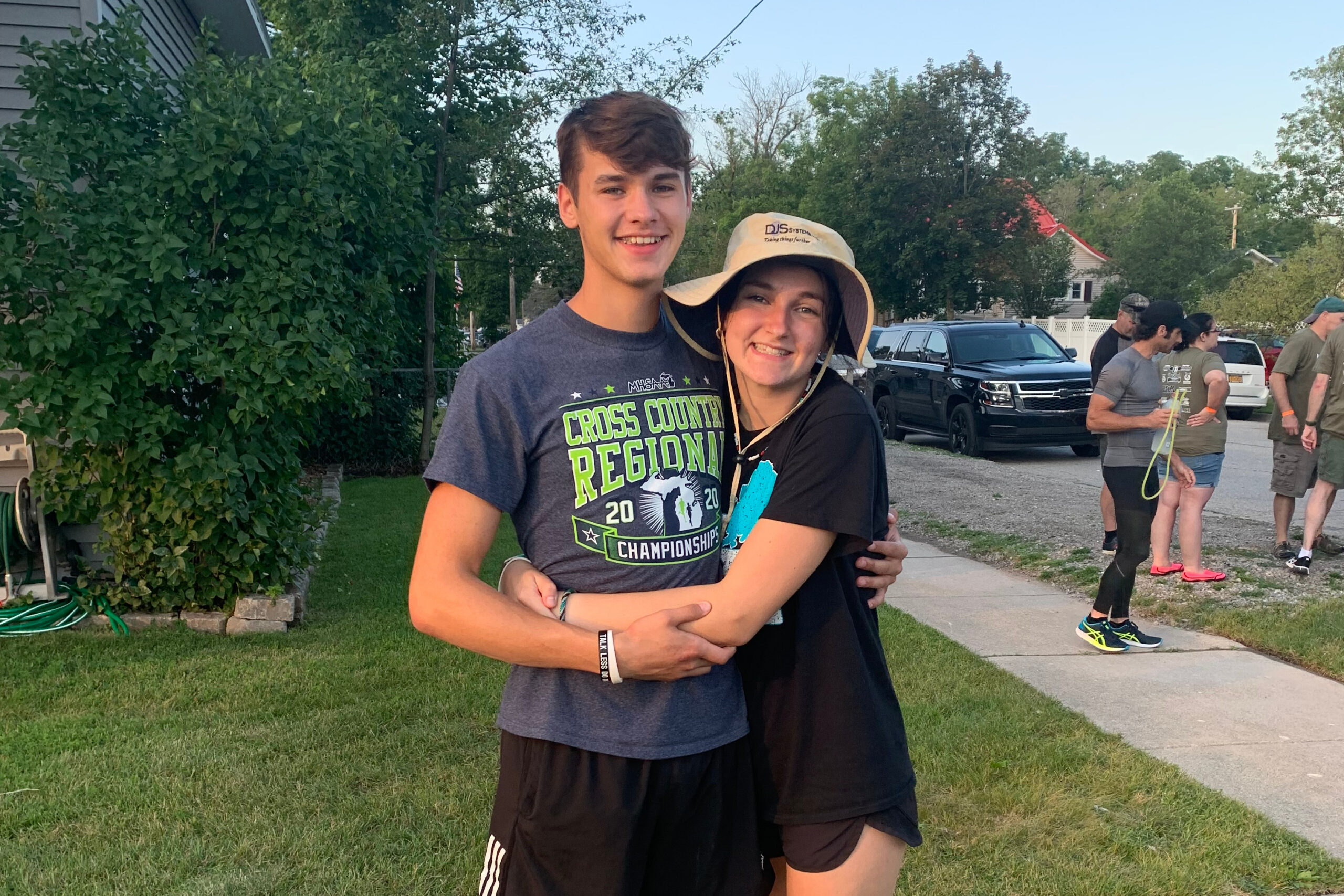 Two smiling teenagers hug in a residential yard during a race event, with volunteers and cars in the background.