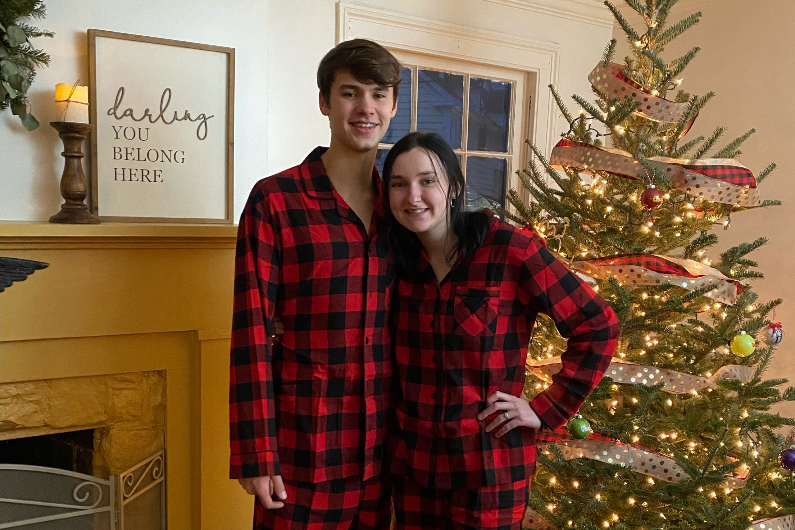 Couple in matching red plaid outfits posing by a Christmas tree and fireplace indoors, smiling at the camera.