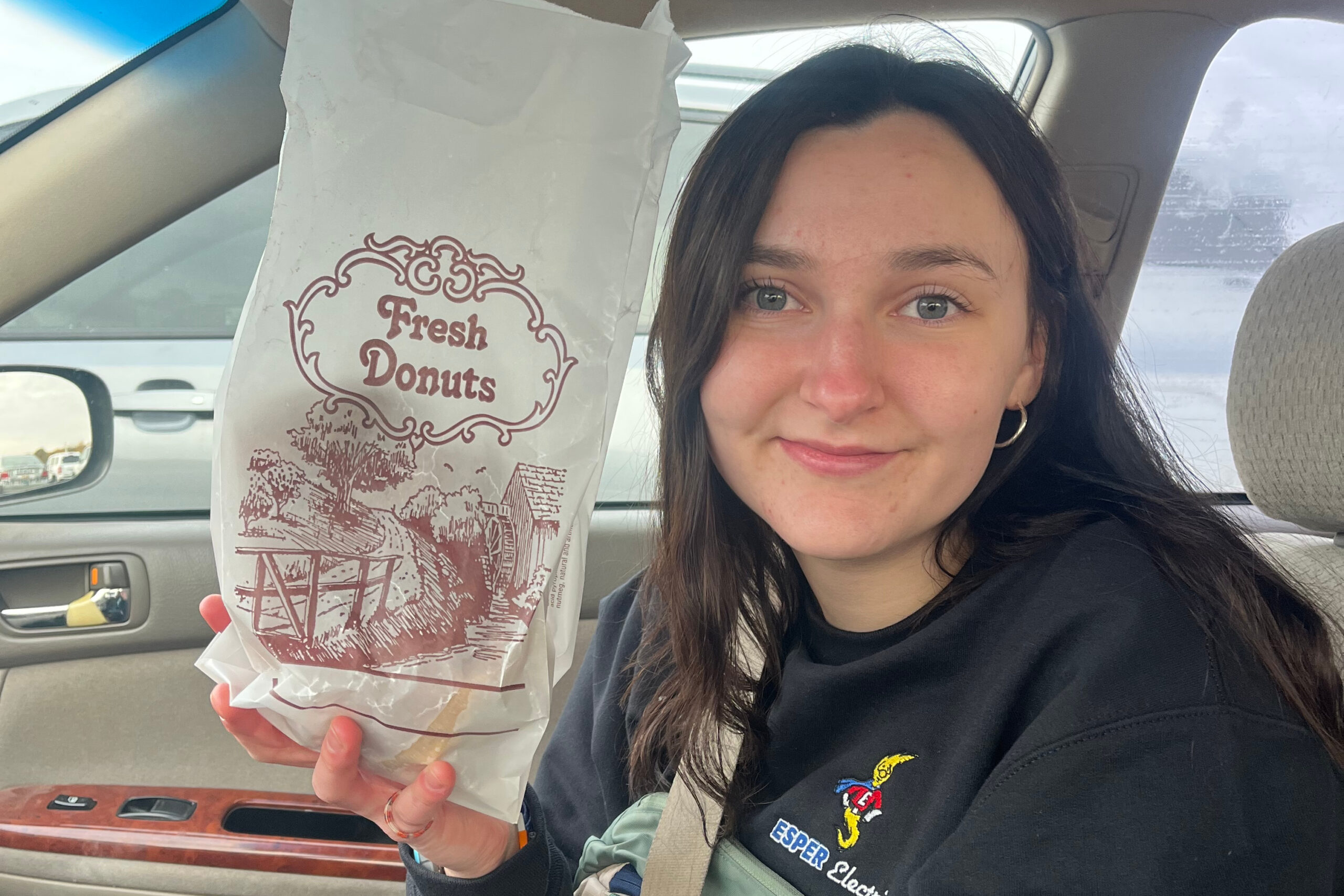 Person in a car holding a Fresh Donuts bag toward the camera, showing the bakery’s logo on the bag