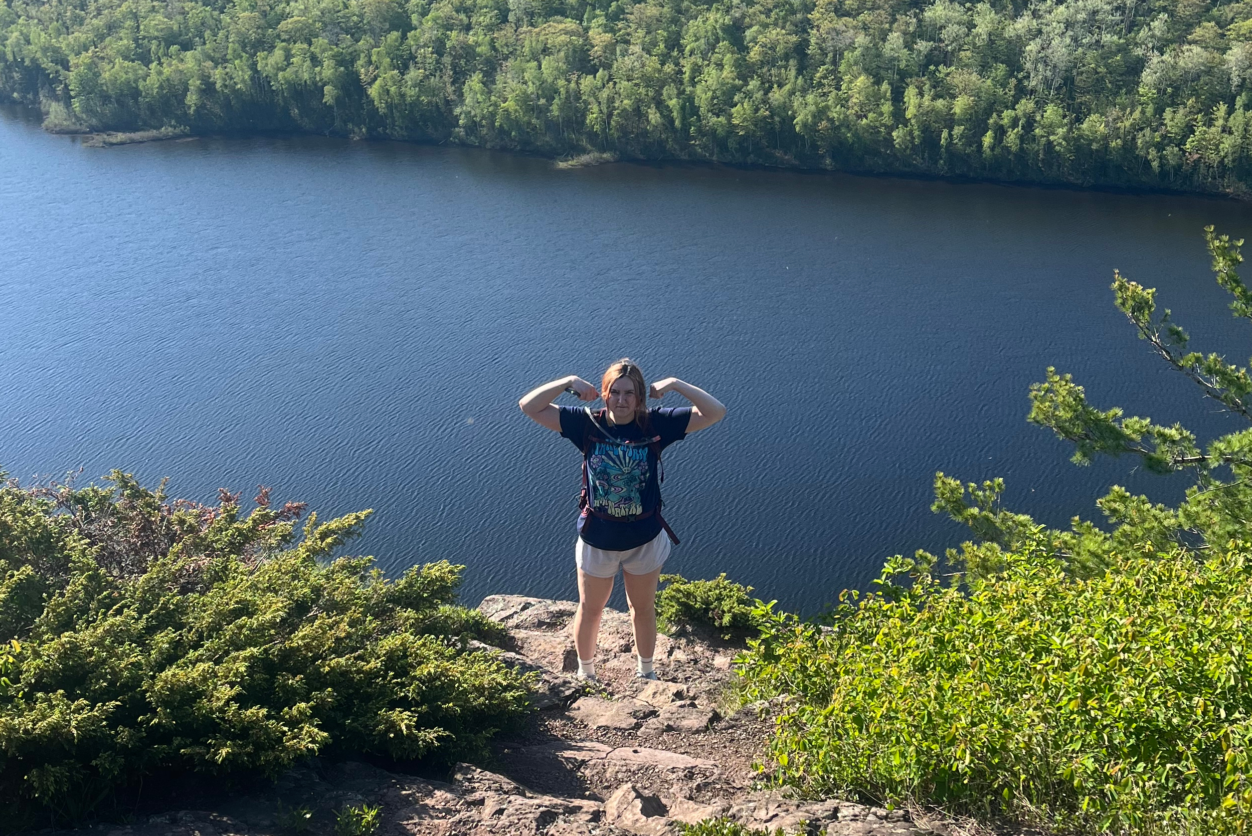 Person standing on a rocky ledge, flexing arms, overlooking a blue lake and dense green forest.
