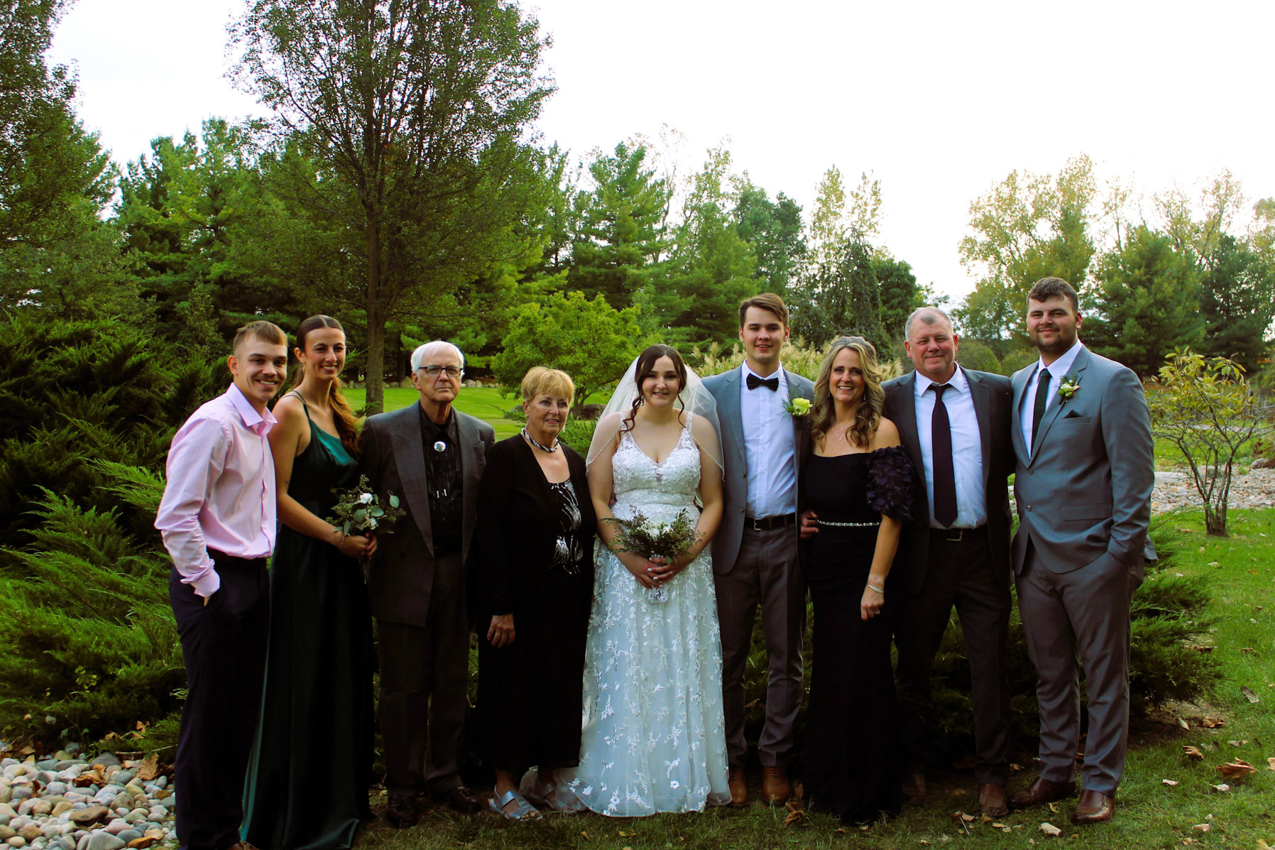 Wedding party posing outdoors on a lawn, bride in a light blue lace gown center, surrounded by family and friends in formal attire.