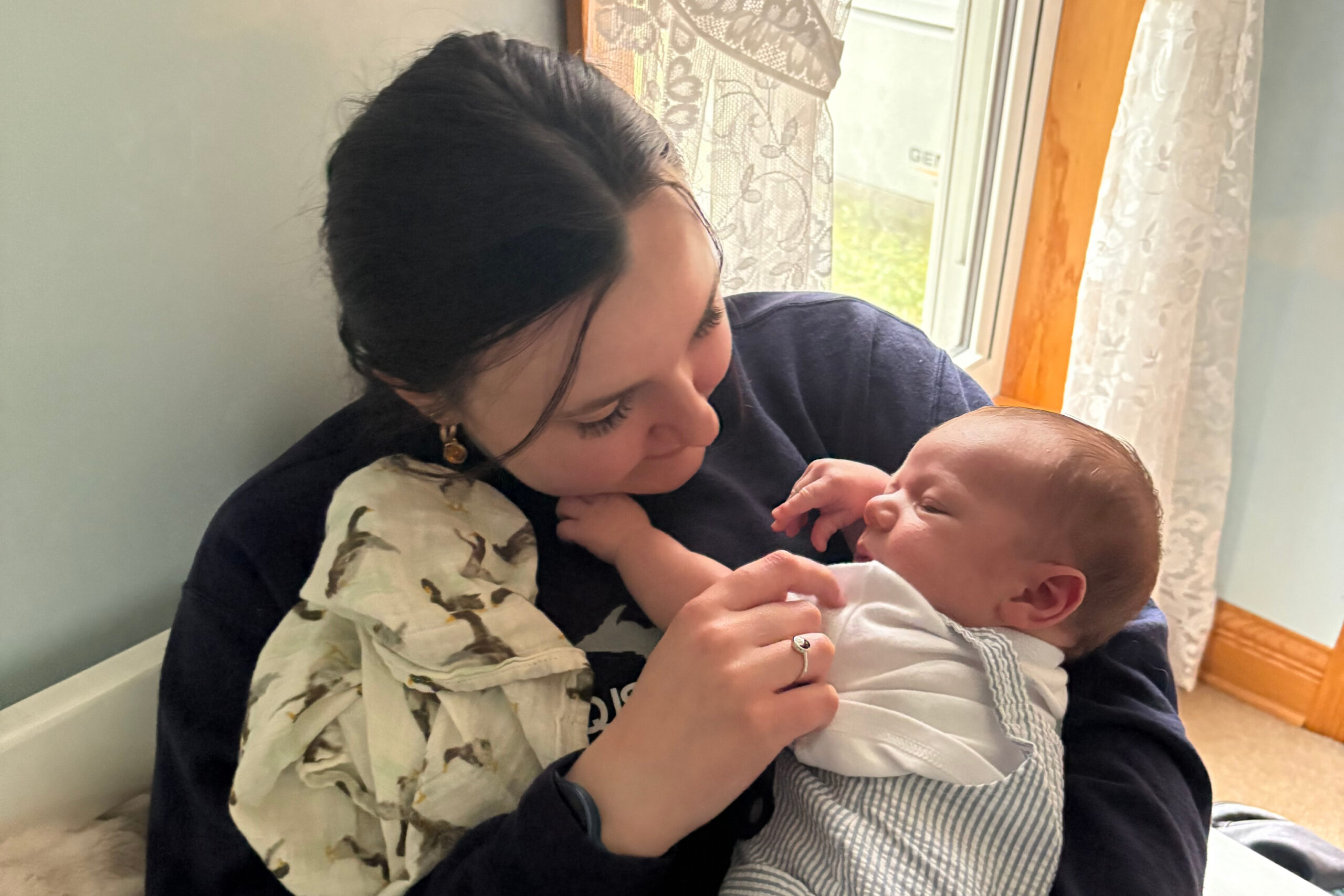 Mother gently holds and kisses her newborn baby near a window with lace curtains.