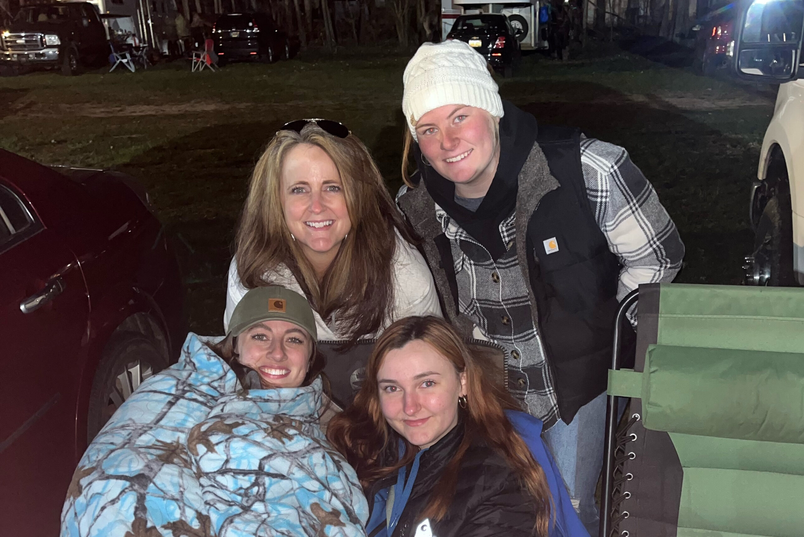 Group of five friends smiling at the camera outdoors at night, wearing jackets and hats and bundled up for cold weather.