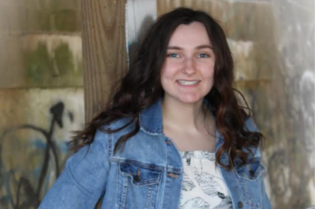 A smiling young woman with long dark hair in a denim jacket stands beside a wooden post outdoors, graffiti-covered wall in the background.