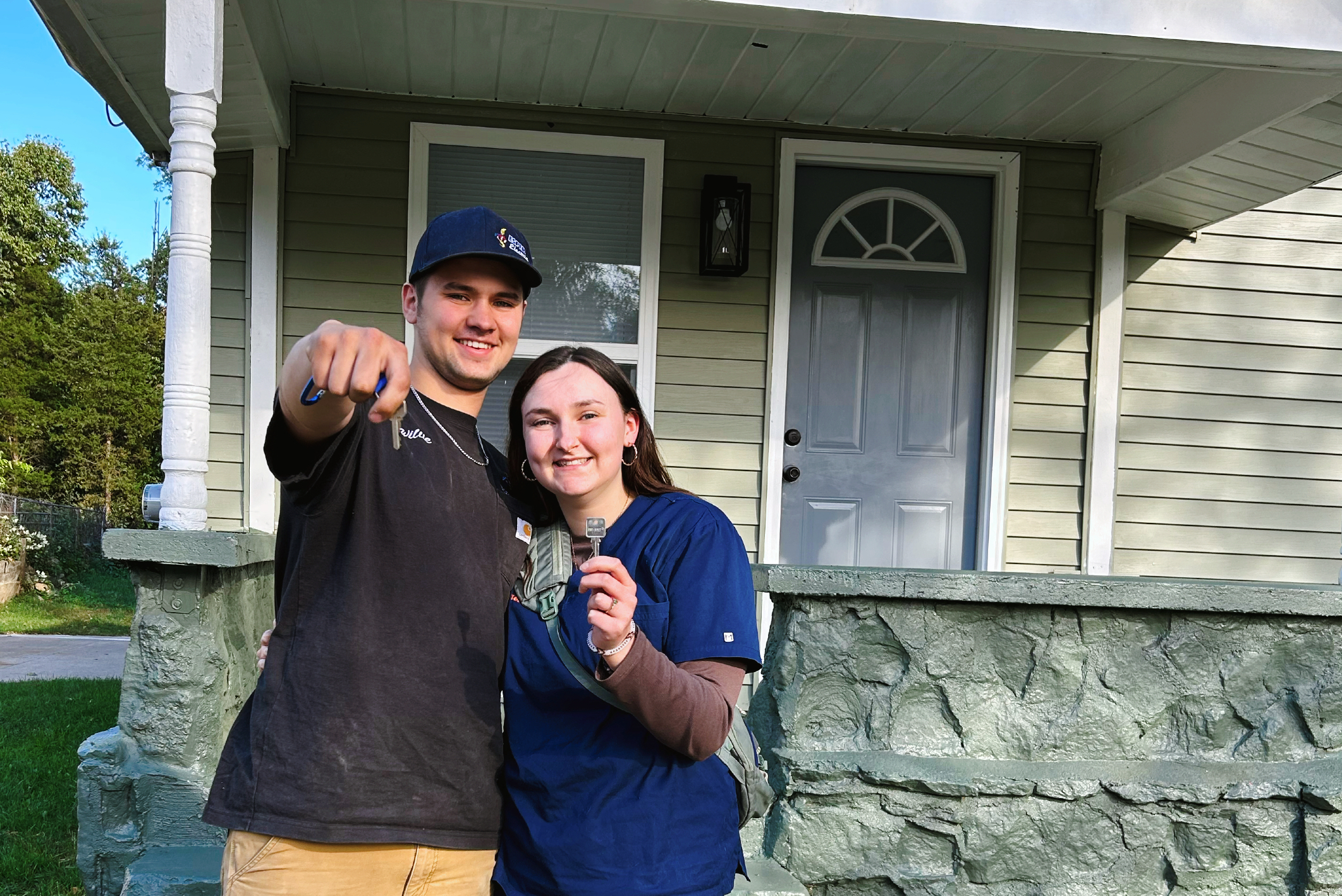 Smiling couple standing on a porch, proudly holding up keys in front of their new home.