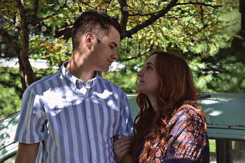Couple stands under a leafy tree, gazing into each other's eyes; the man wears a blue striped shirt and the woman a floral dress, arms linked.