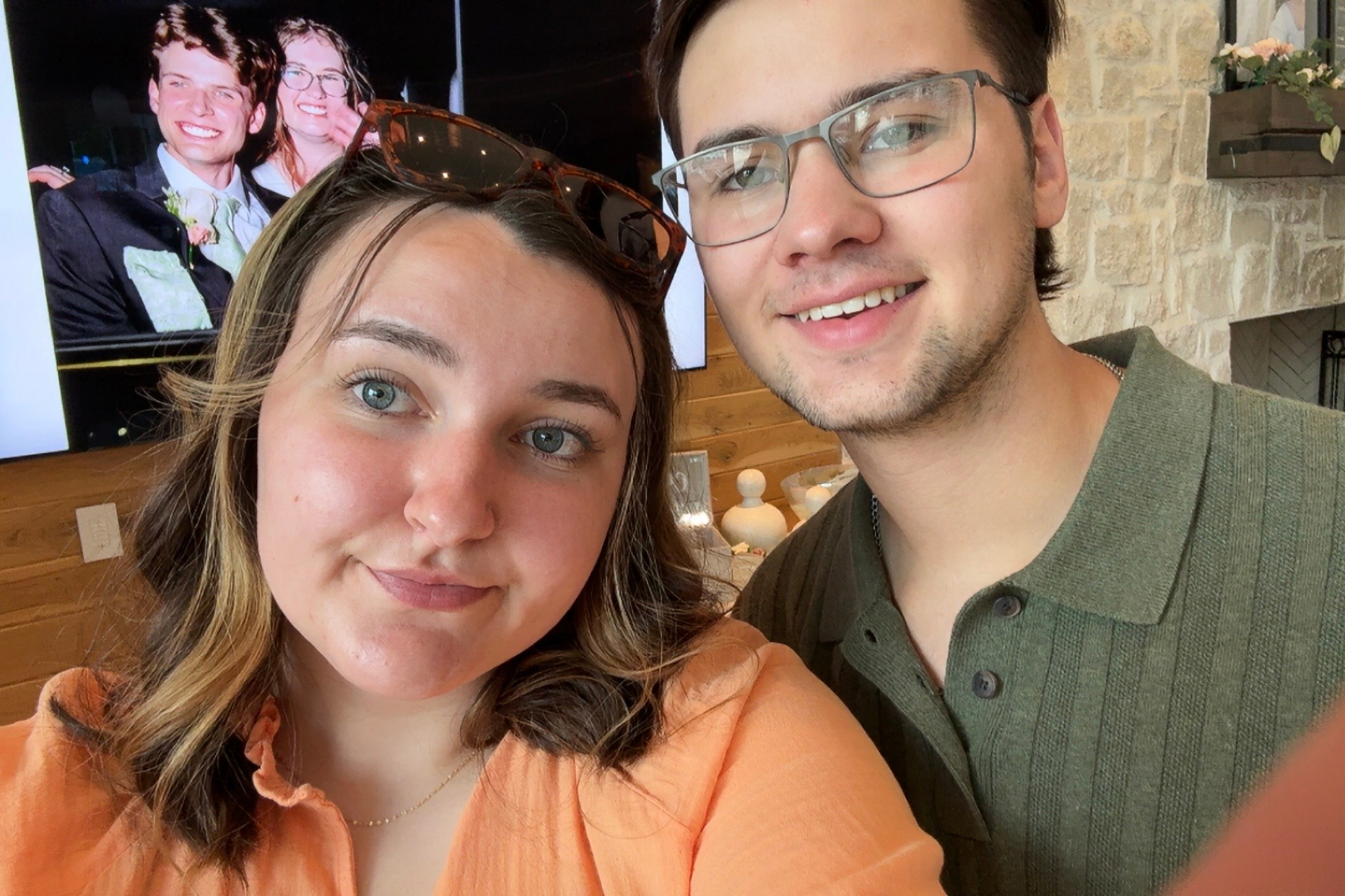 Close-up selfie of a smiling couple indoors: woman in orange top on left, man in green shirt on right, stone wall behind them.