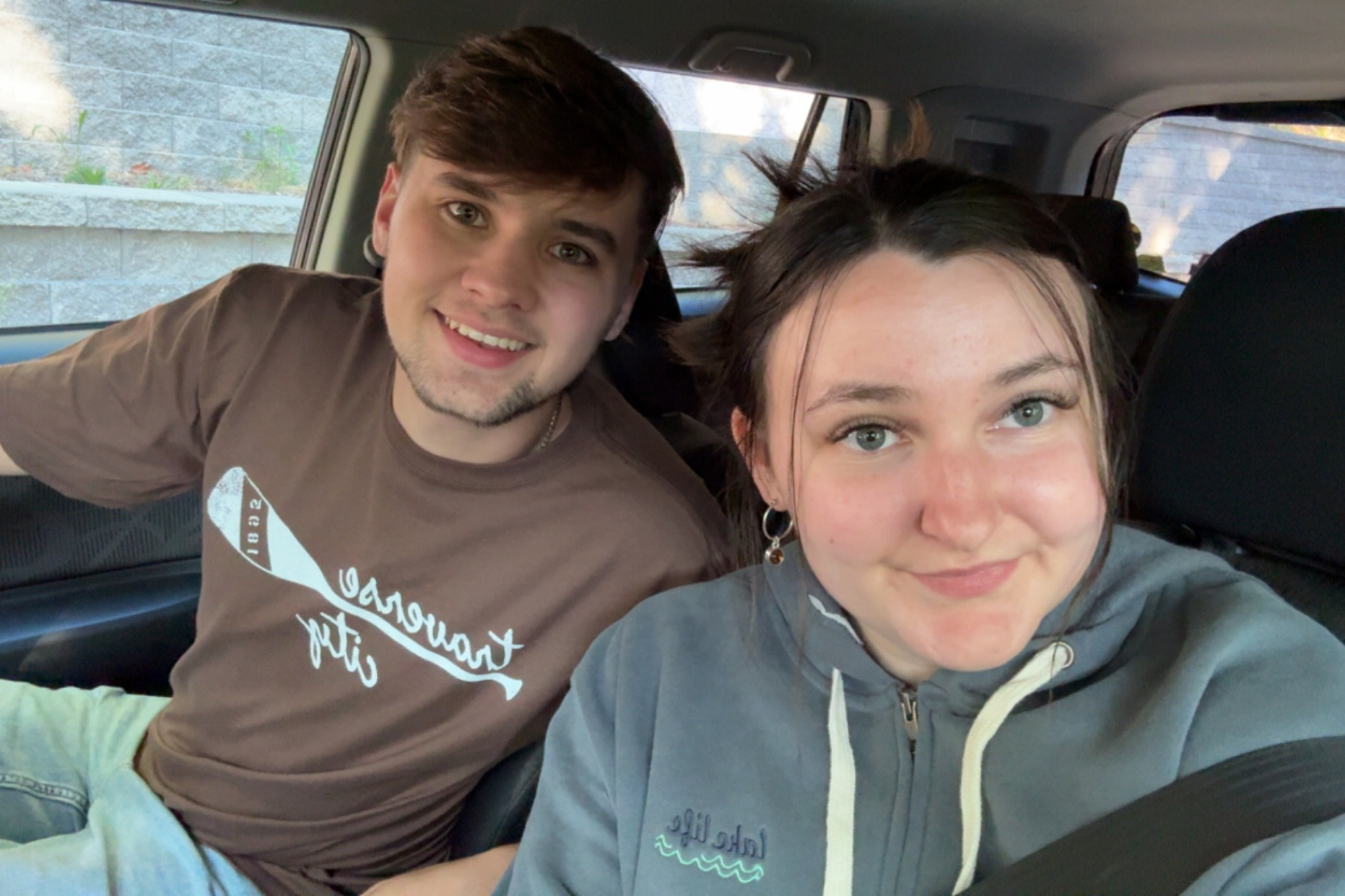 Two friends taking a selfie in a car, smiling at the camera (man on left in brown shirt, woman on right in gray hoodie).