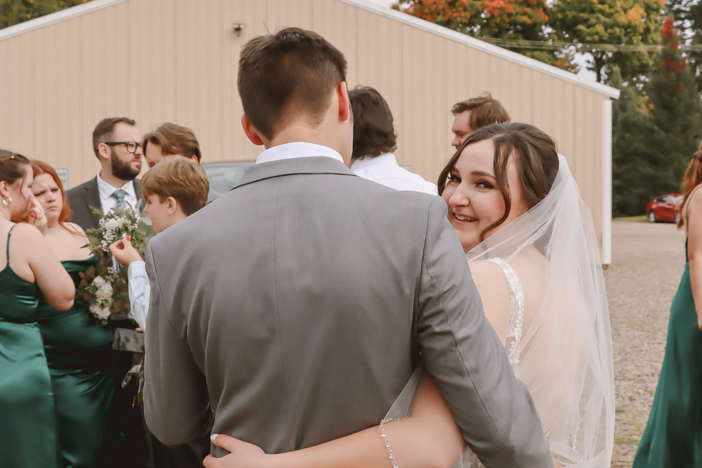 Bride in veil hugging groom in a gray suit at an outdoor wedding, smiling at the camera, with guests in green dresses behind them.