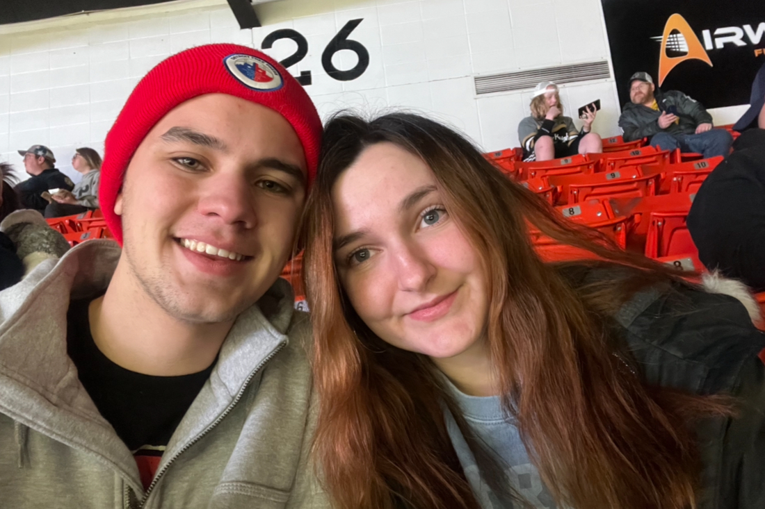 Selfie of a smiling couple in a stadium with red seats; man wearing a red knit beanie and gray jacket beside a woman with long hair.