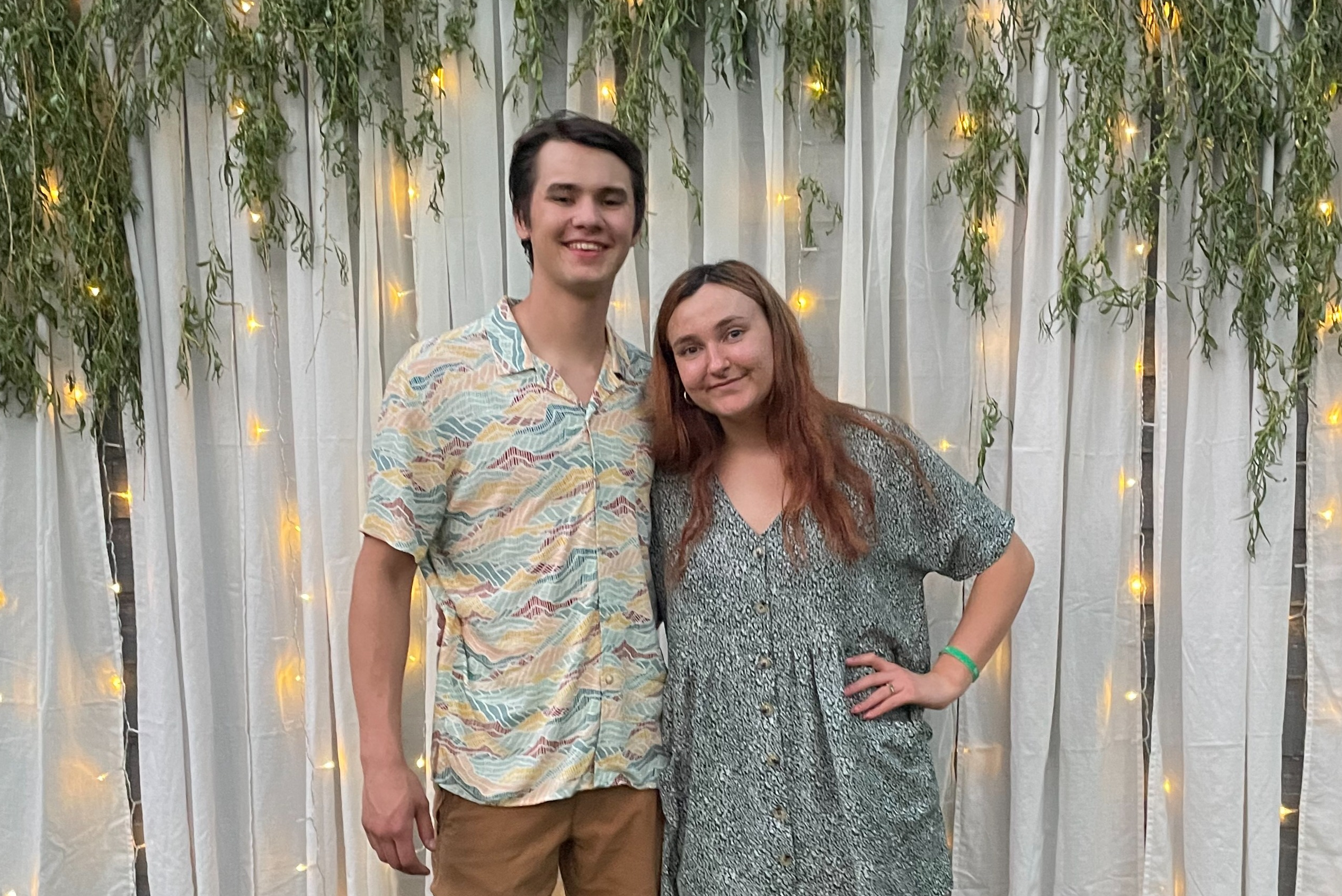 Two people pose together in front of a white curtain backdrop with hanging greenery and warm string lights at an event.Paraphrase: A couple smiles at the camera in front of decorative lights and vines.