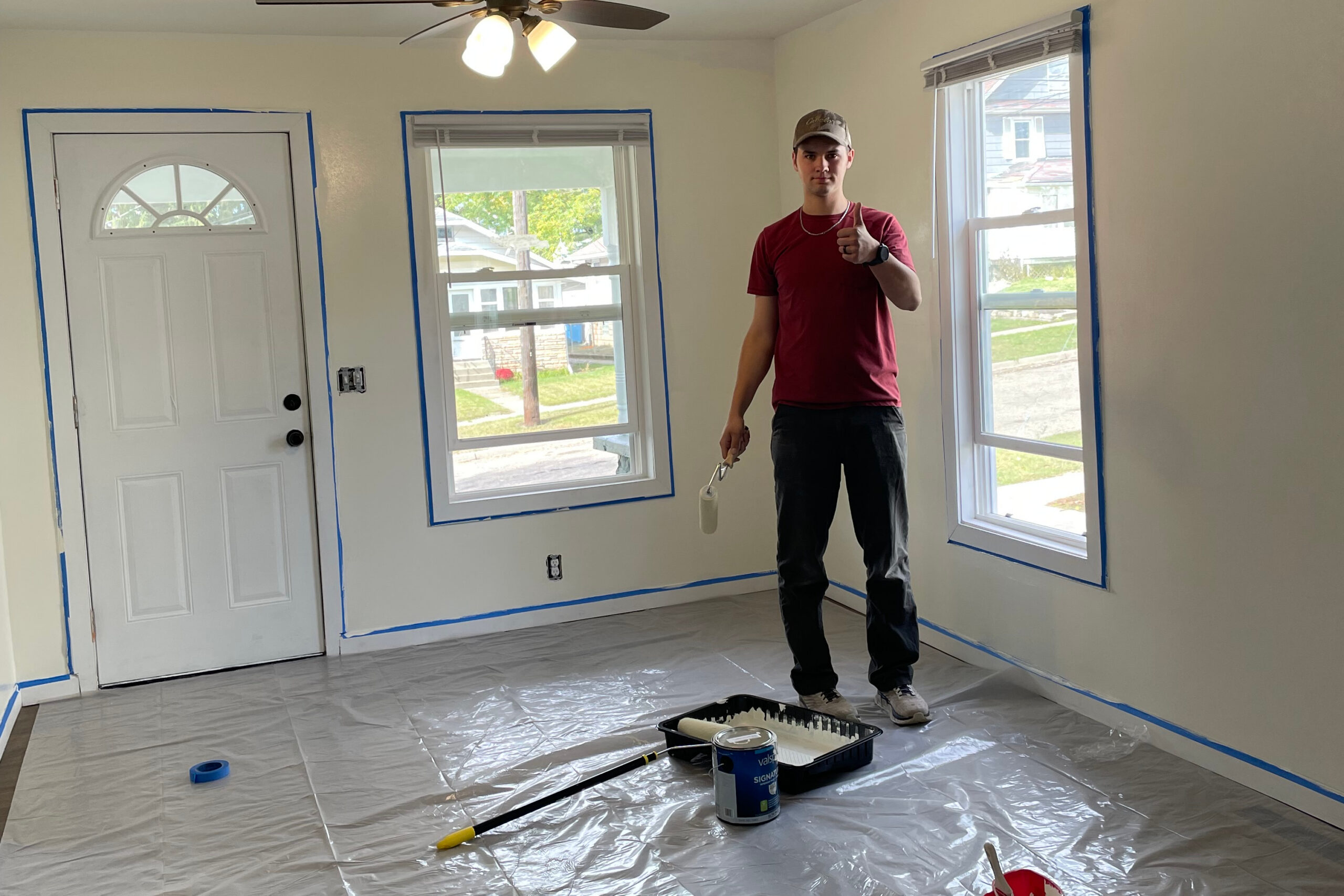 Person in a red shirt stands indoors giving a thumbs-up while painting a room; painter's tape around windows and door, plastic-covered floor, paint tray and rollers nearby.