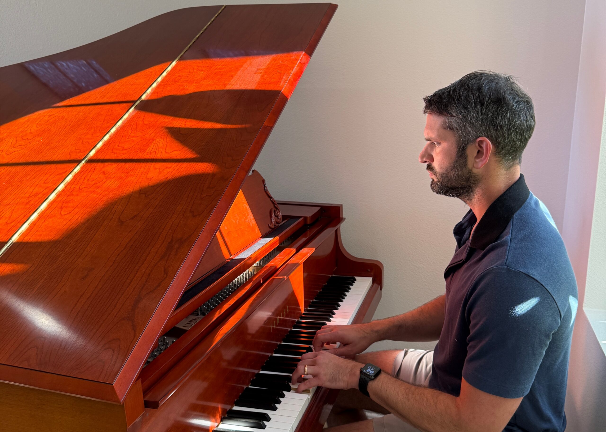 Man wearing a dark polo plays a glossy wooden upright piano, hands on the keyboard and focus on playing.