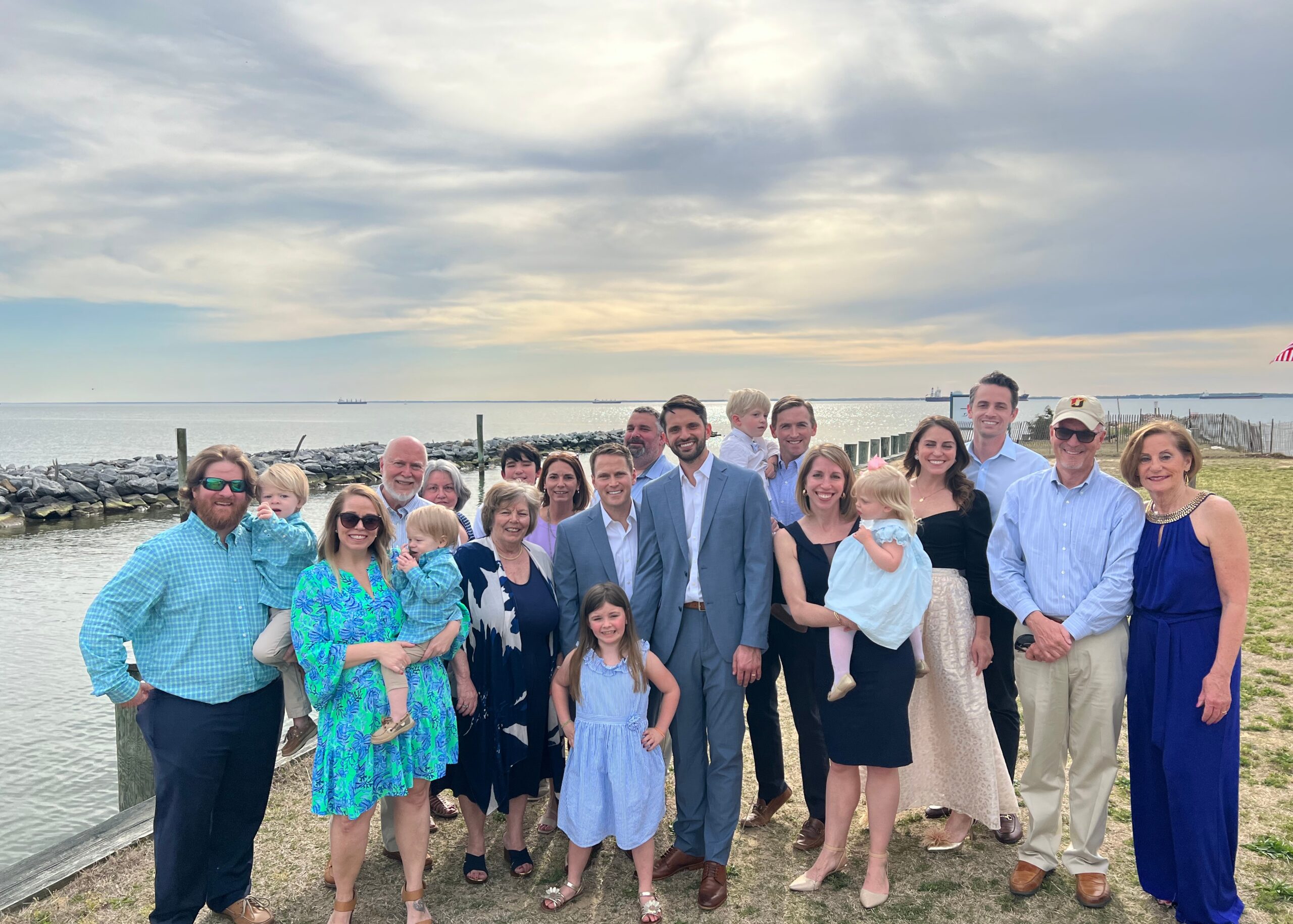 Large group of family and friends posing together on a seaside boardwalk at dusk, smiling.
