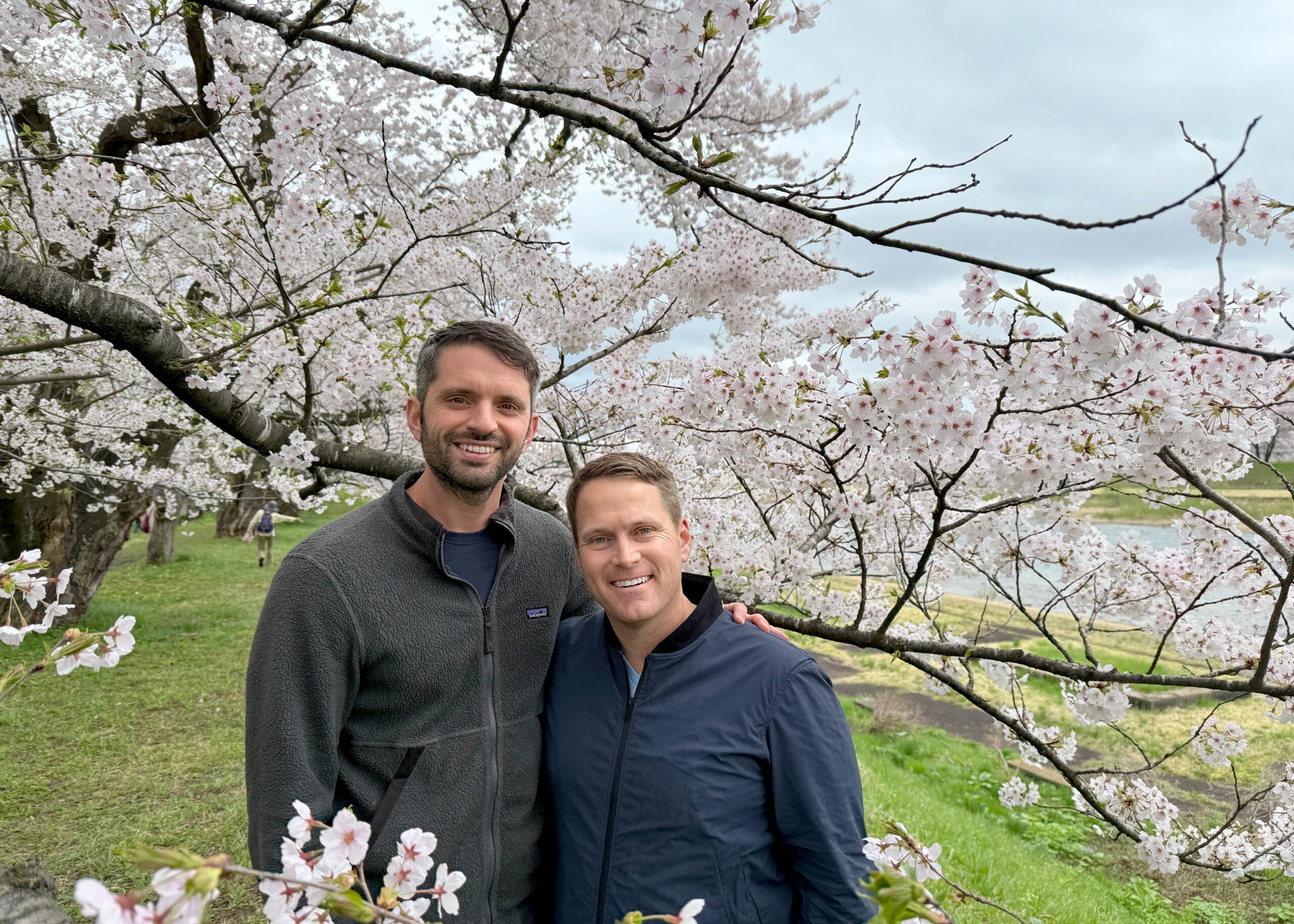 Two men smiling and posing for a photo beneath cherry blossom branches in a park with green grass behind them.