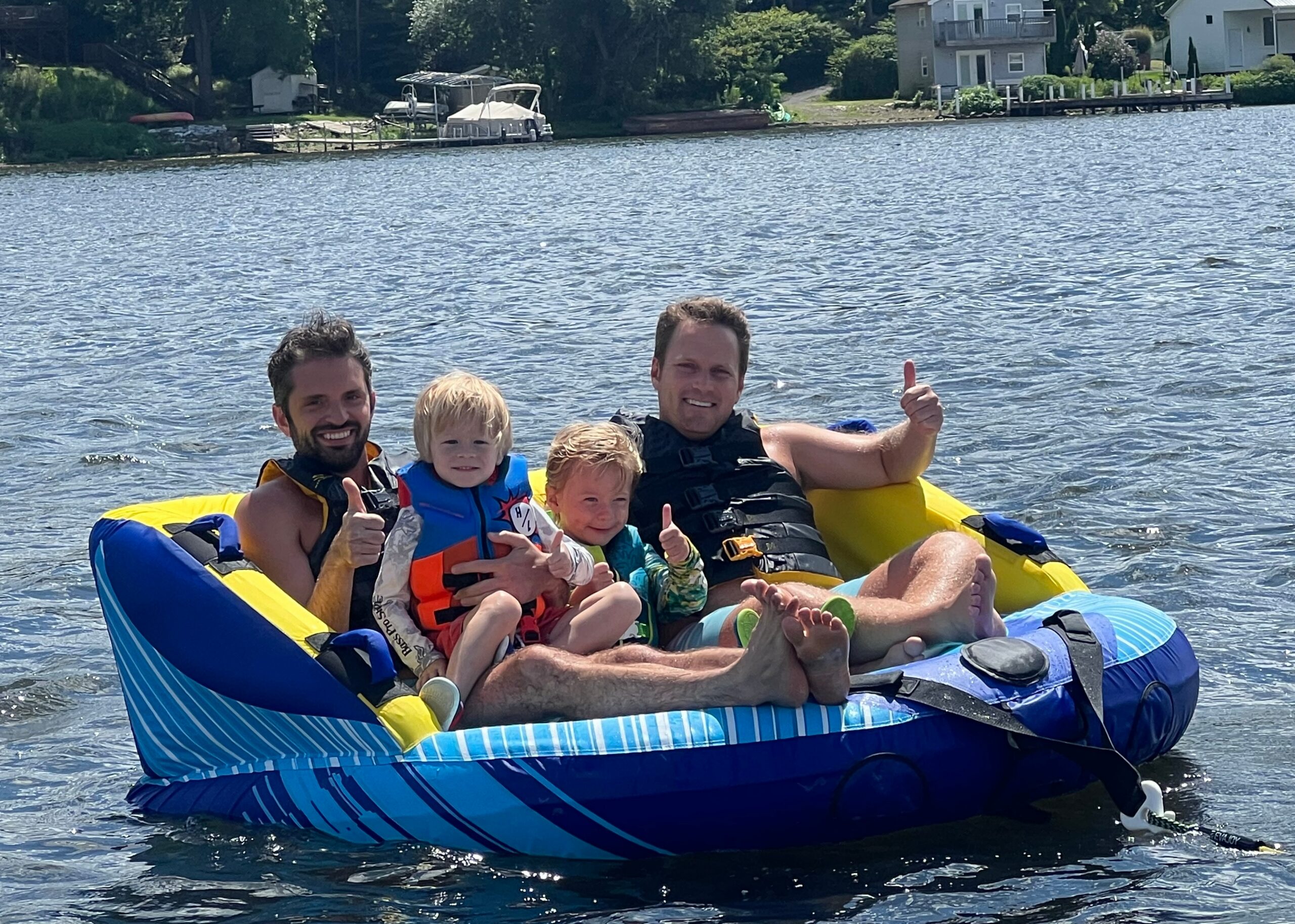Family of four on a blue inflatable raft giving thumbs up on a lake, wearing life jackets, sunny day near houses on shore