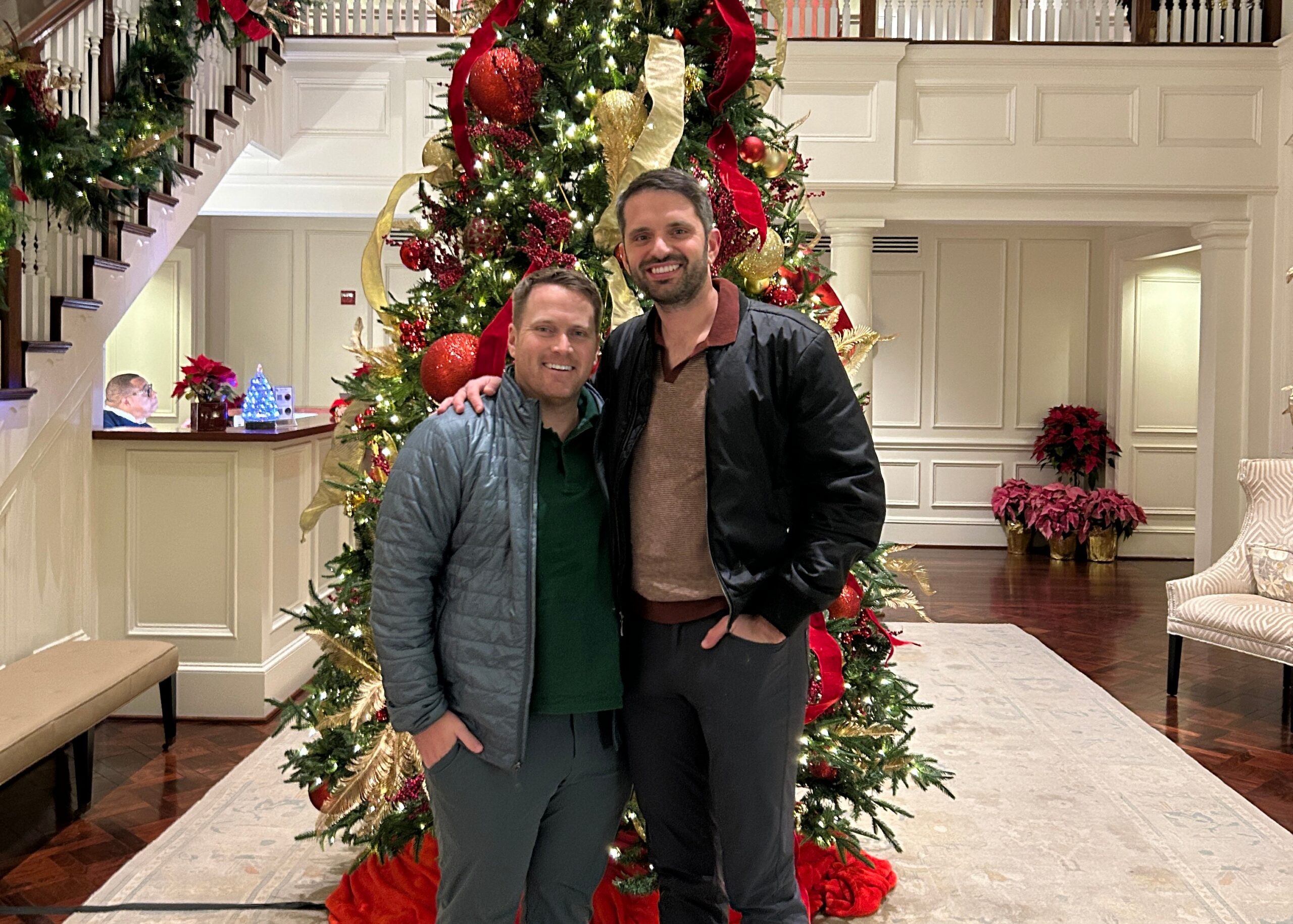 Two men posing together in a hotel lobby next to an elaborately decorated Christmas tree with red and gold ornaments and ribbons.