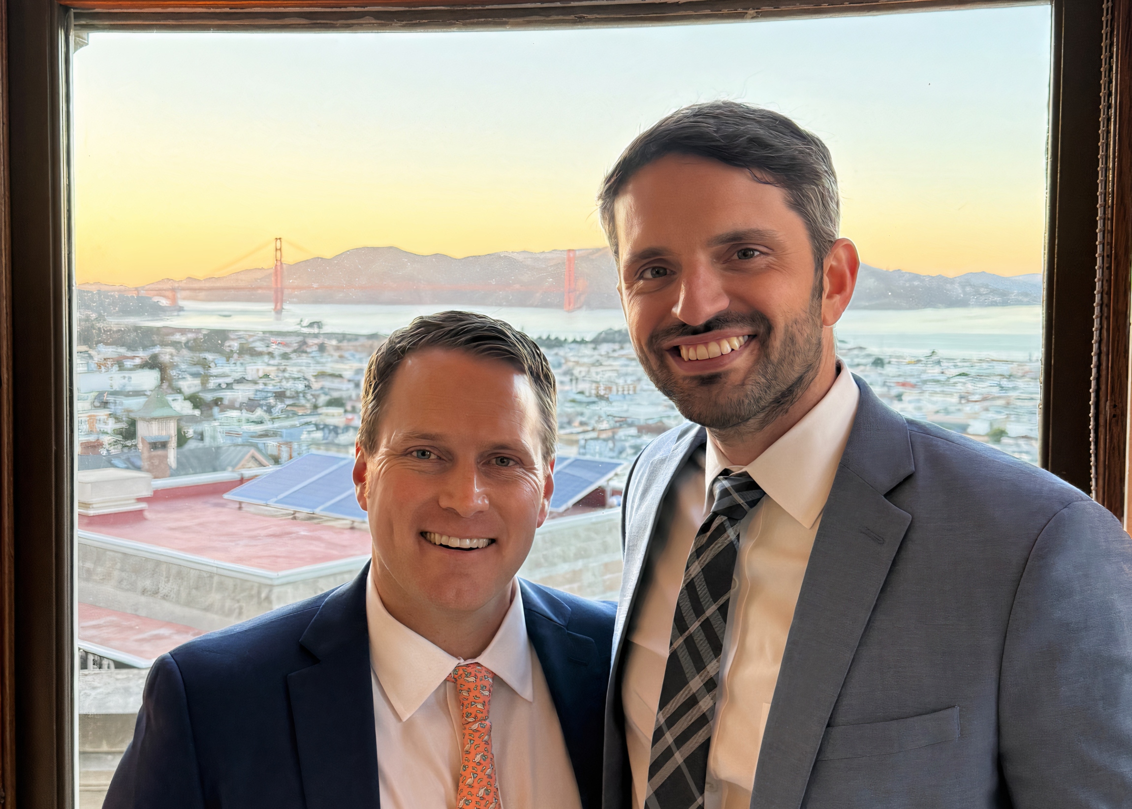 Two men in suits smiling for a selfie in front of a window overlooking a city with the Golden Gate Bridge at sunset in the background, visible between them, creating a cheerful group portrait on a trip.