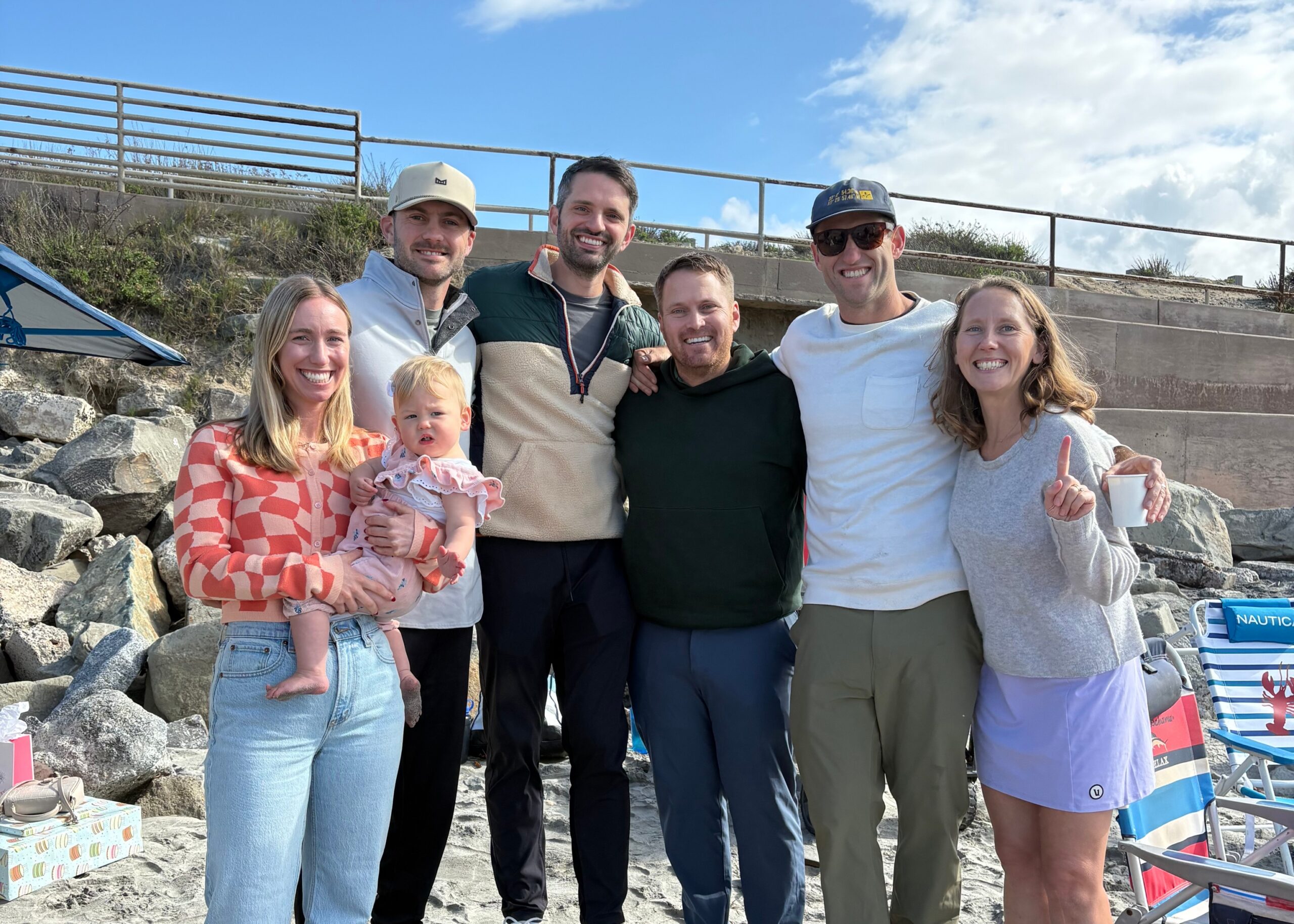 Group of six adults and a baby posing on a rocky beach with a blue sky backdrop, smiling.