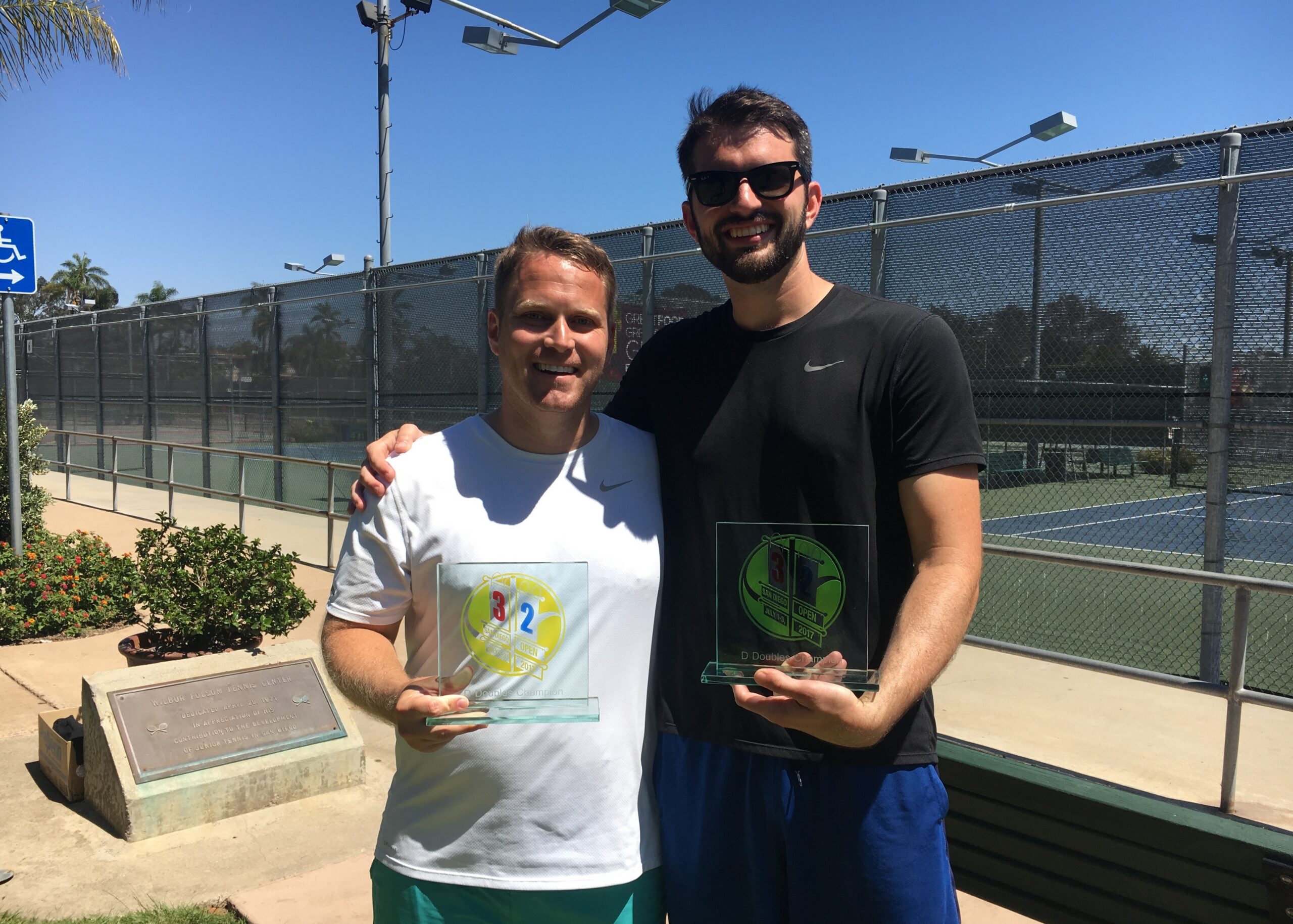 Two men stand together outdoors after a tennis match, each holding glass trophies and smiling at the camera with a chain-link fence and court in the background.