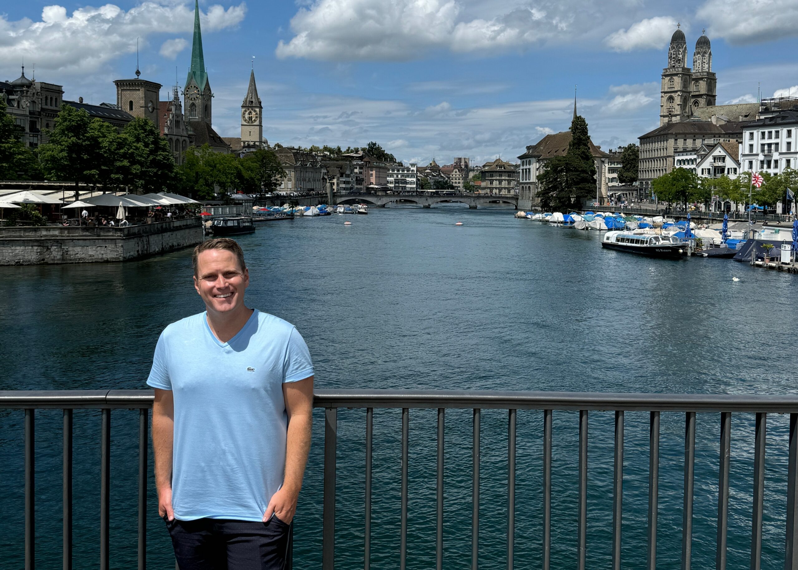 Man in a light blue shirt standing at a railing along a river, with a historic cityscape and boats in the background.
