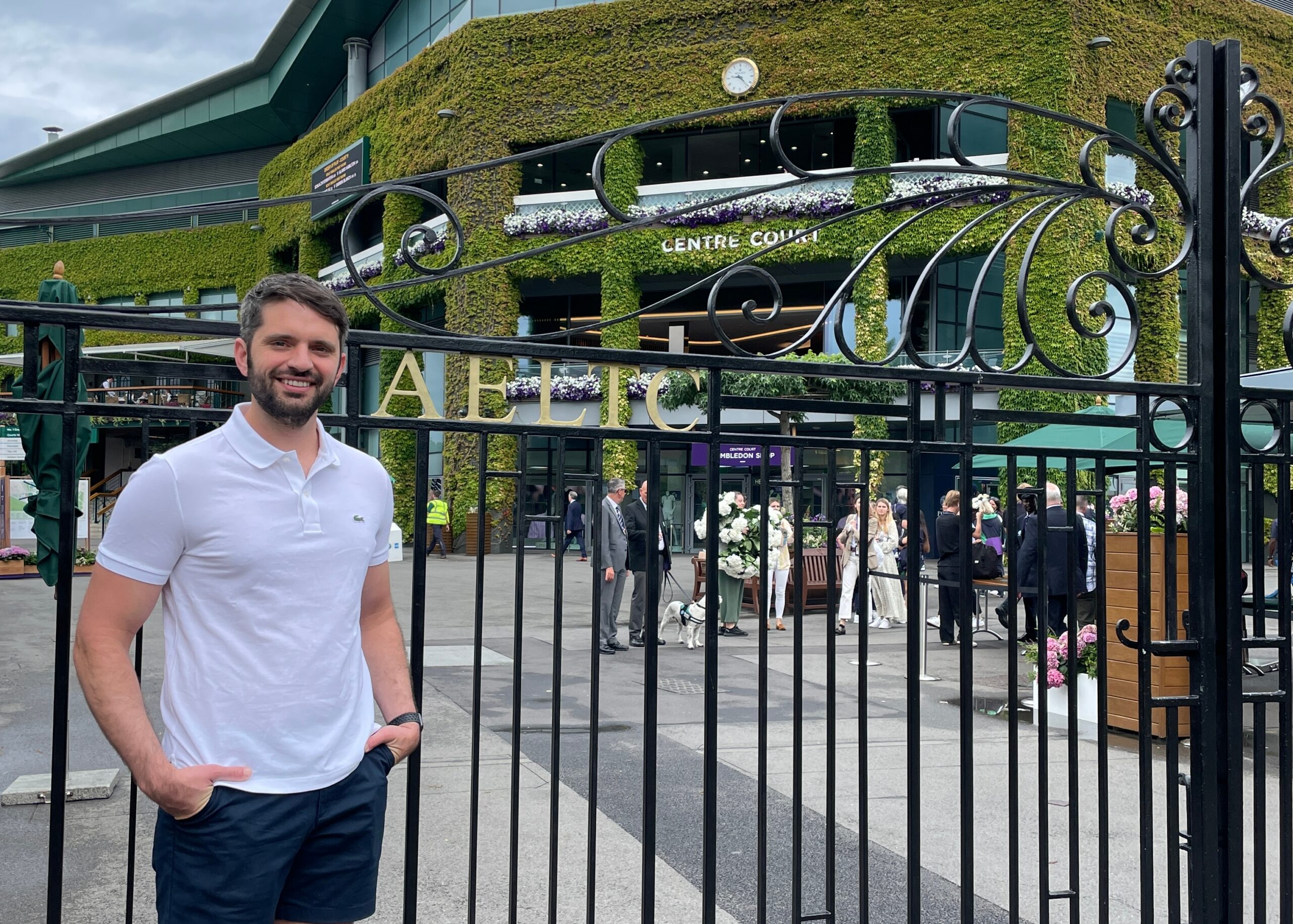 Man in a white polo shirt standing beside a decorative black gate outside a green ivy-covered building with people in formal attire in the background.