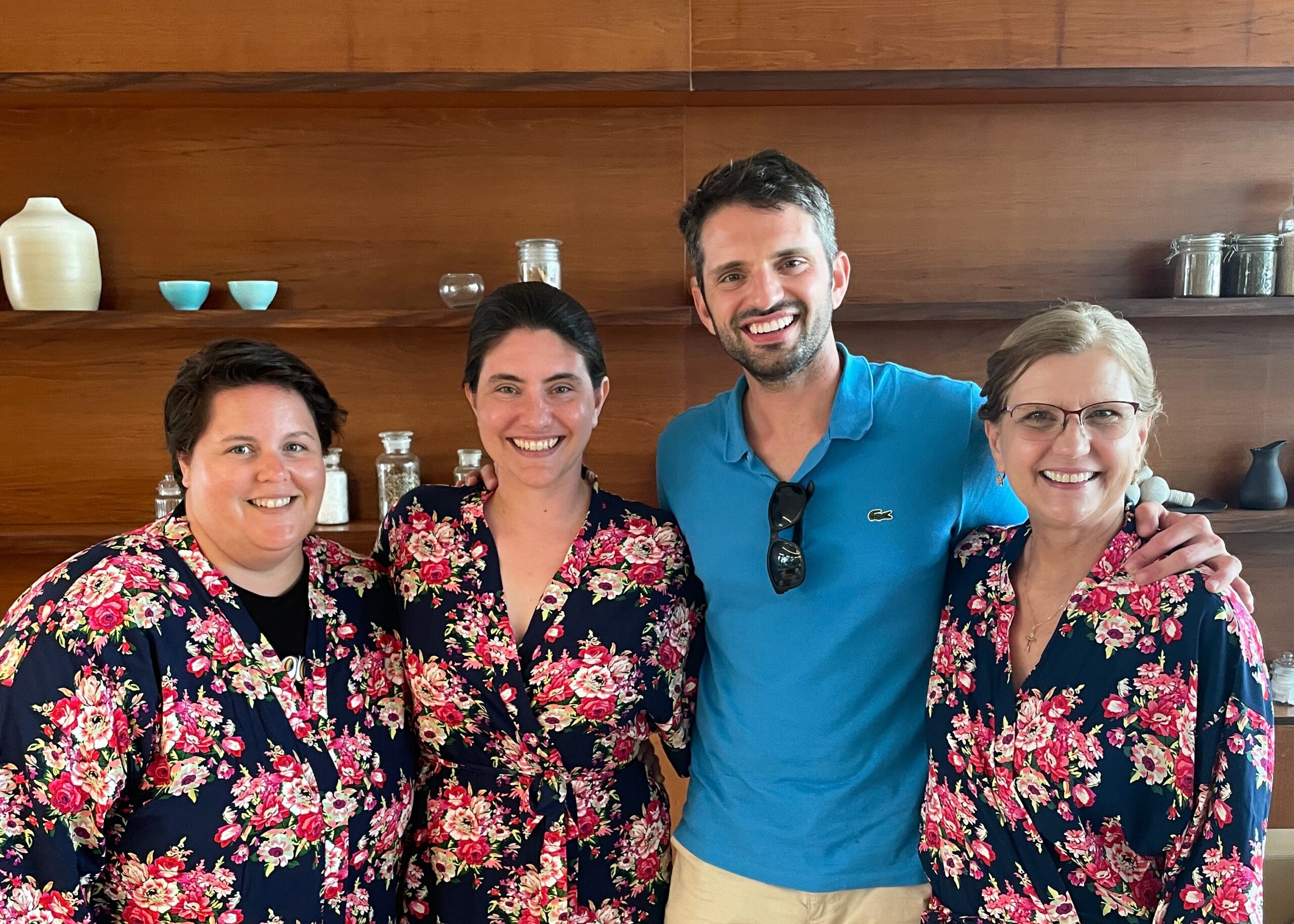 Four adults smiling and posing together, three women in navy floral dresses and a man in a blue polo, indoors against a wood-paneled backdrop.
