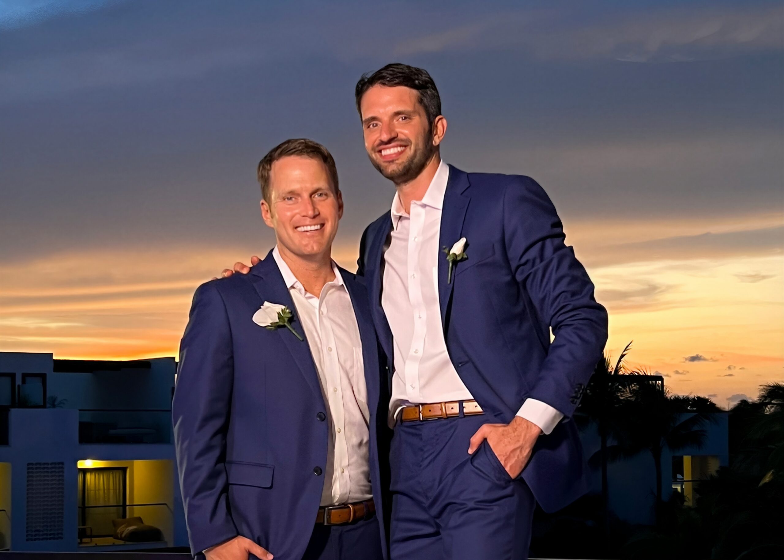 Two men in blue suits posing together at sunset, smiling with arms around each other.