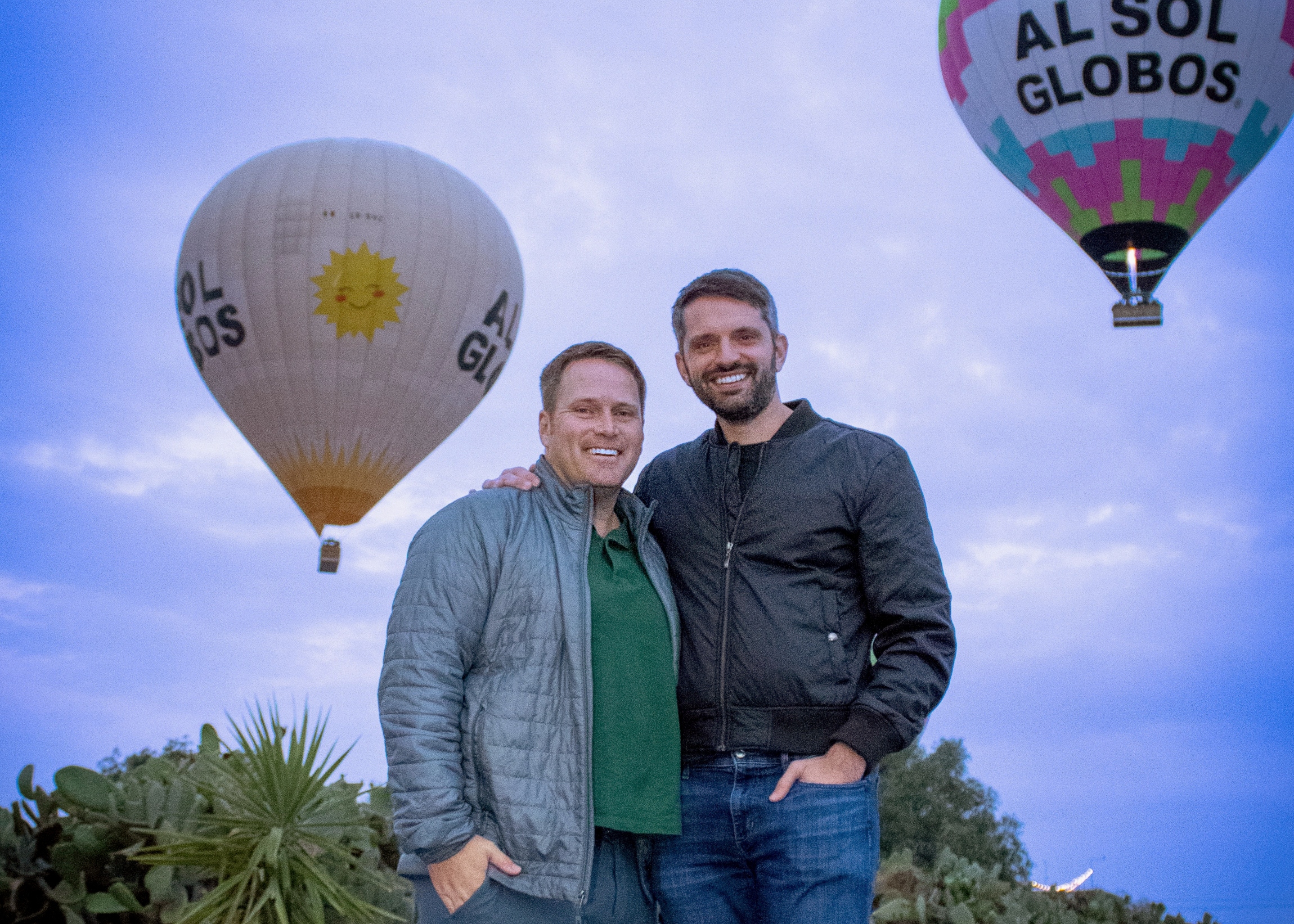 Two men standing close together outdoors, with colorful hot air balloons in the blue sky behind them.